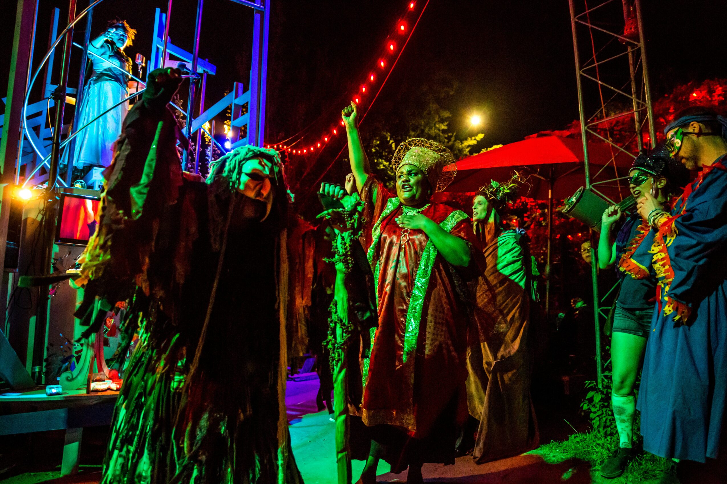 Group of performers in colorful costumes and masks on stage at night, with bright lights and audience nearby.