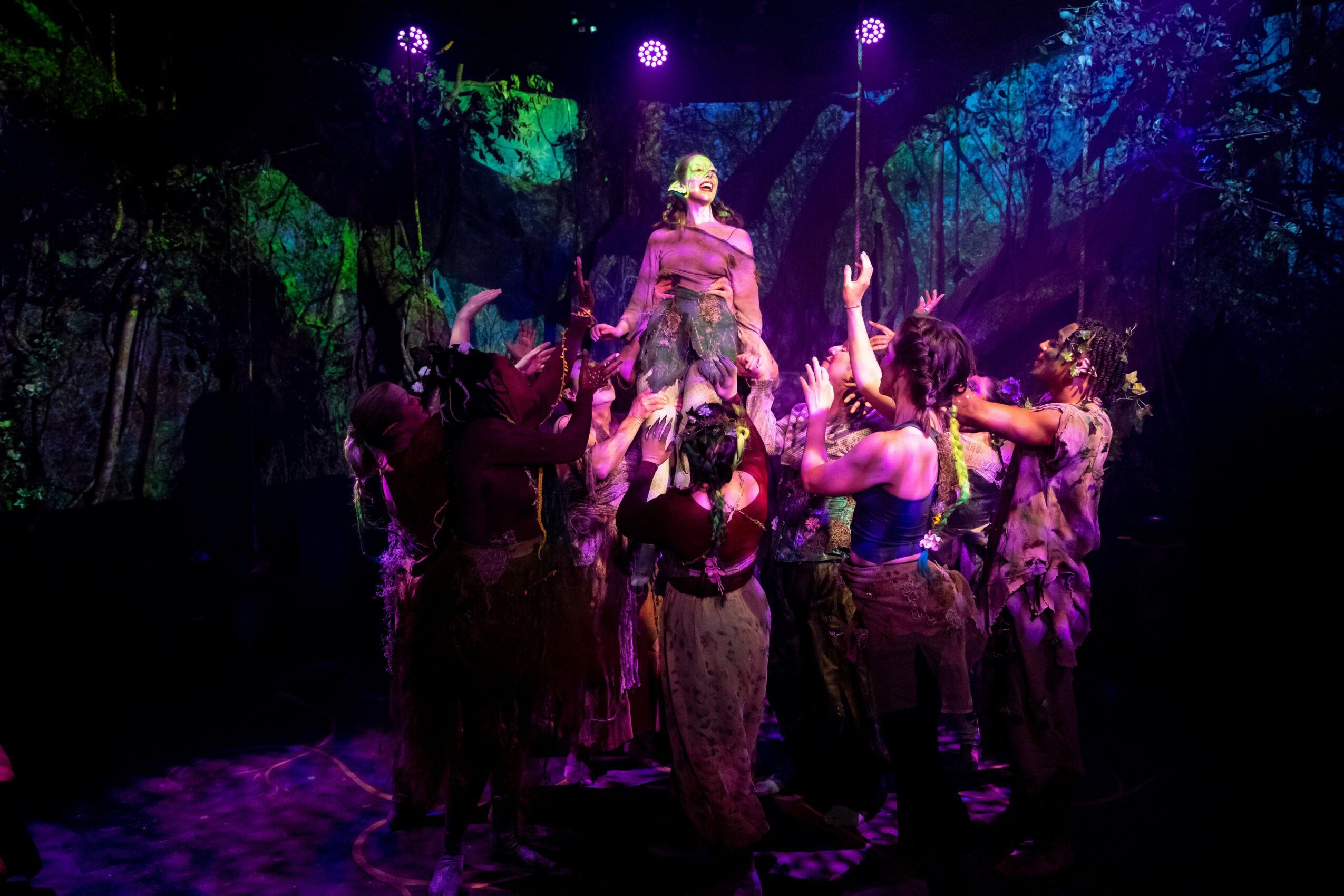 Group of performers on stage with colorful lighting, some reaching upward, in a theatrical scene.