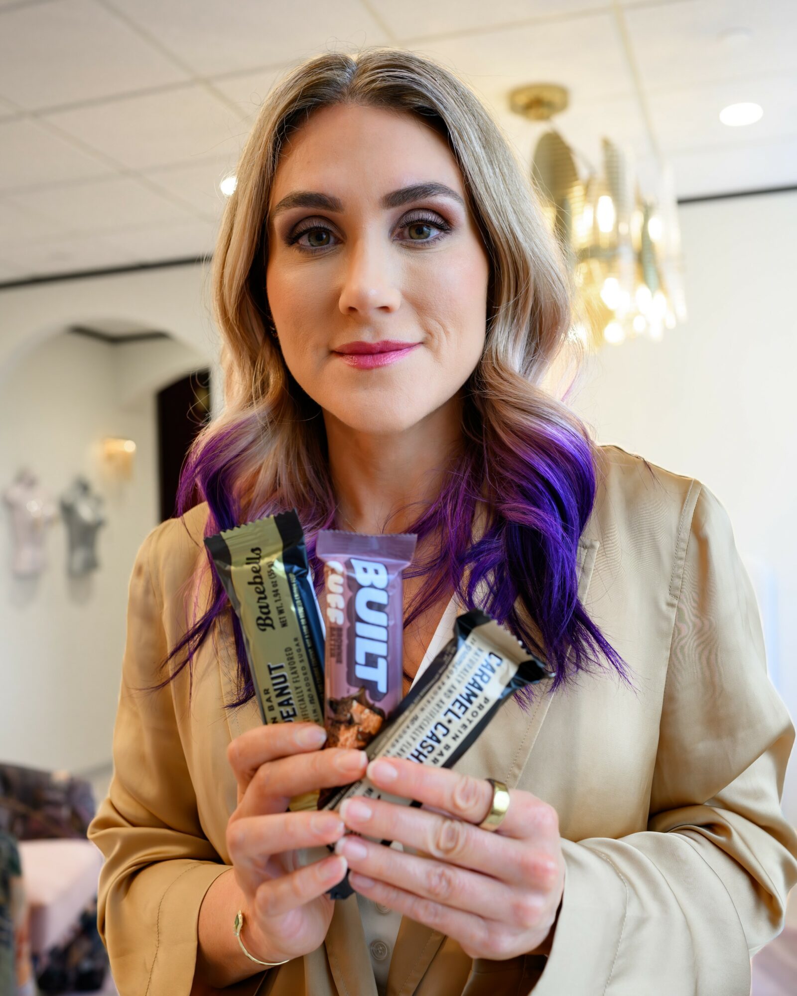 Young woman with wavy hair, purple tips, holding multiple snack bars, smiling in an indoor setting with chandelier and decor.