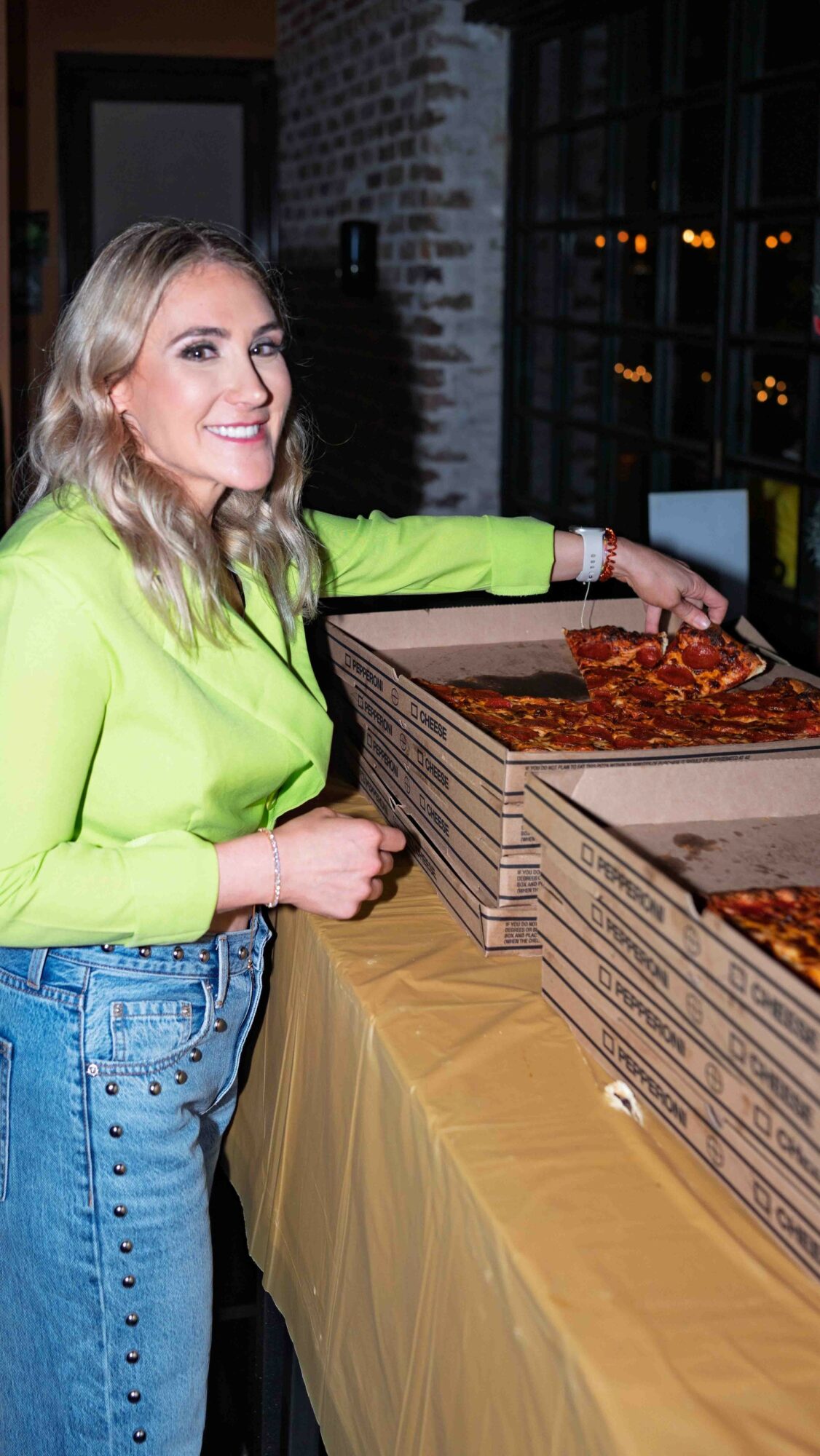 Woman in neon green top smiling and reaching for pizza boxes on table, brick wall and window in background.