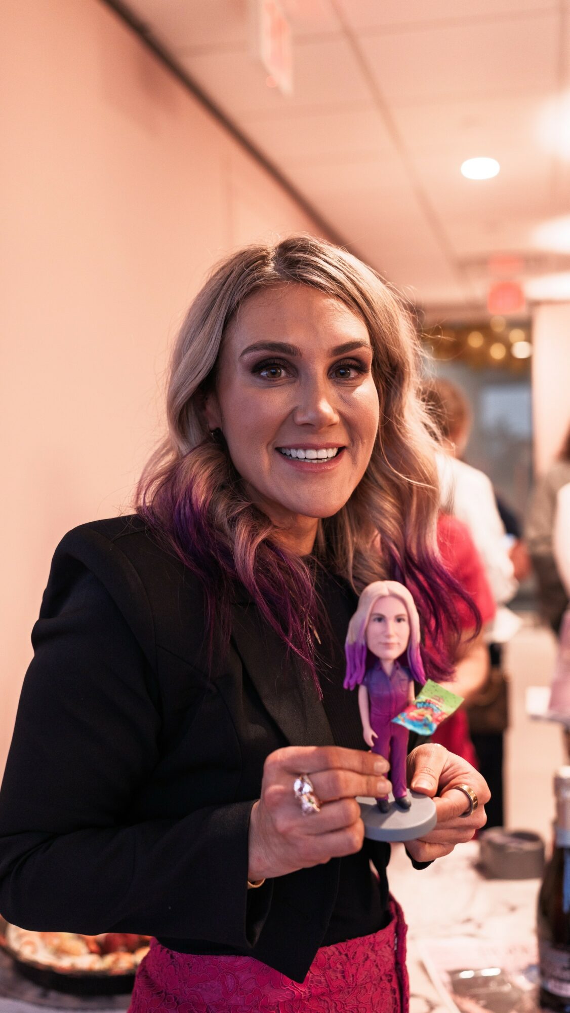 Woman with blonde and purple hair smiling, holding a small doll, in an indoor setting.