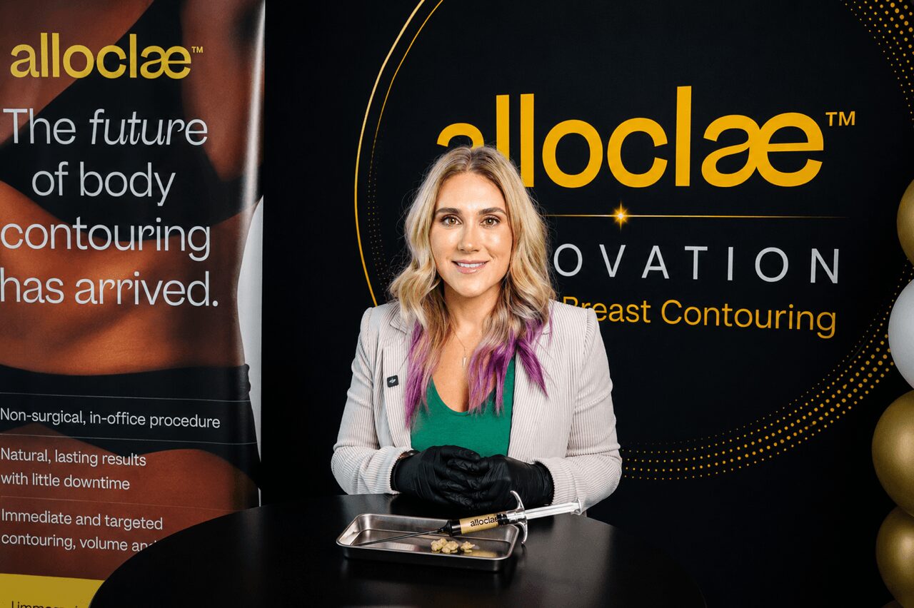 Woman with blonde hair in a white blazer and purple shirt sitting at a table, smiling, in front of a black and gold backdrop with text.