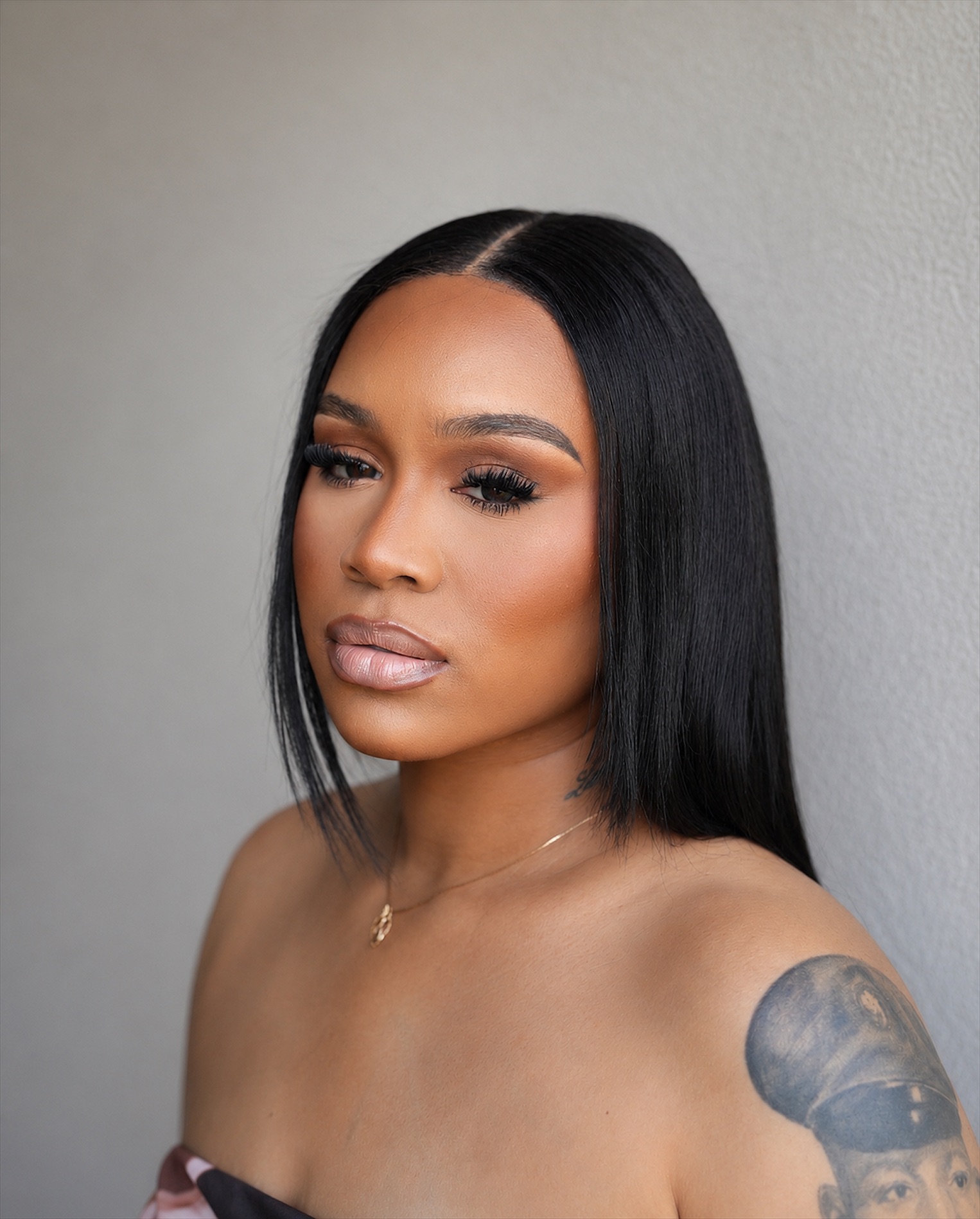 Portrait of a woman with straight black hair, makeup, and a tattoo on her shoulder, against a plain wall.