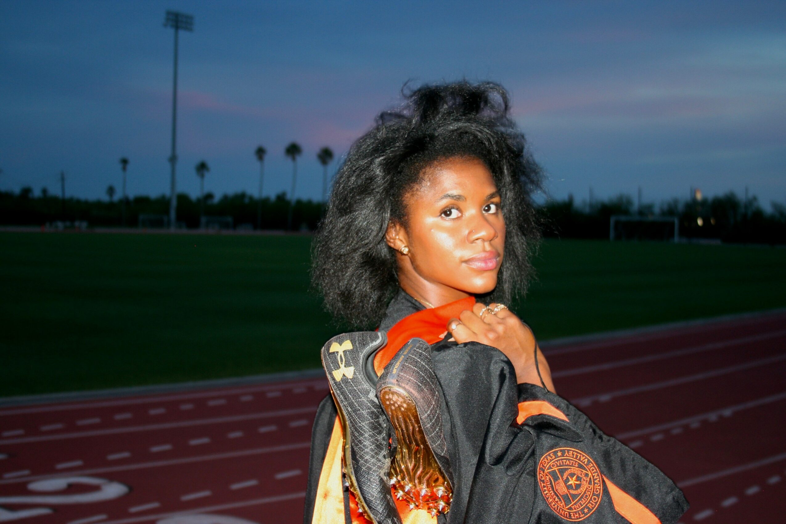 Young woman with natural hair standing on a running track at dusk, looking at the camera, with a sports field in the background.