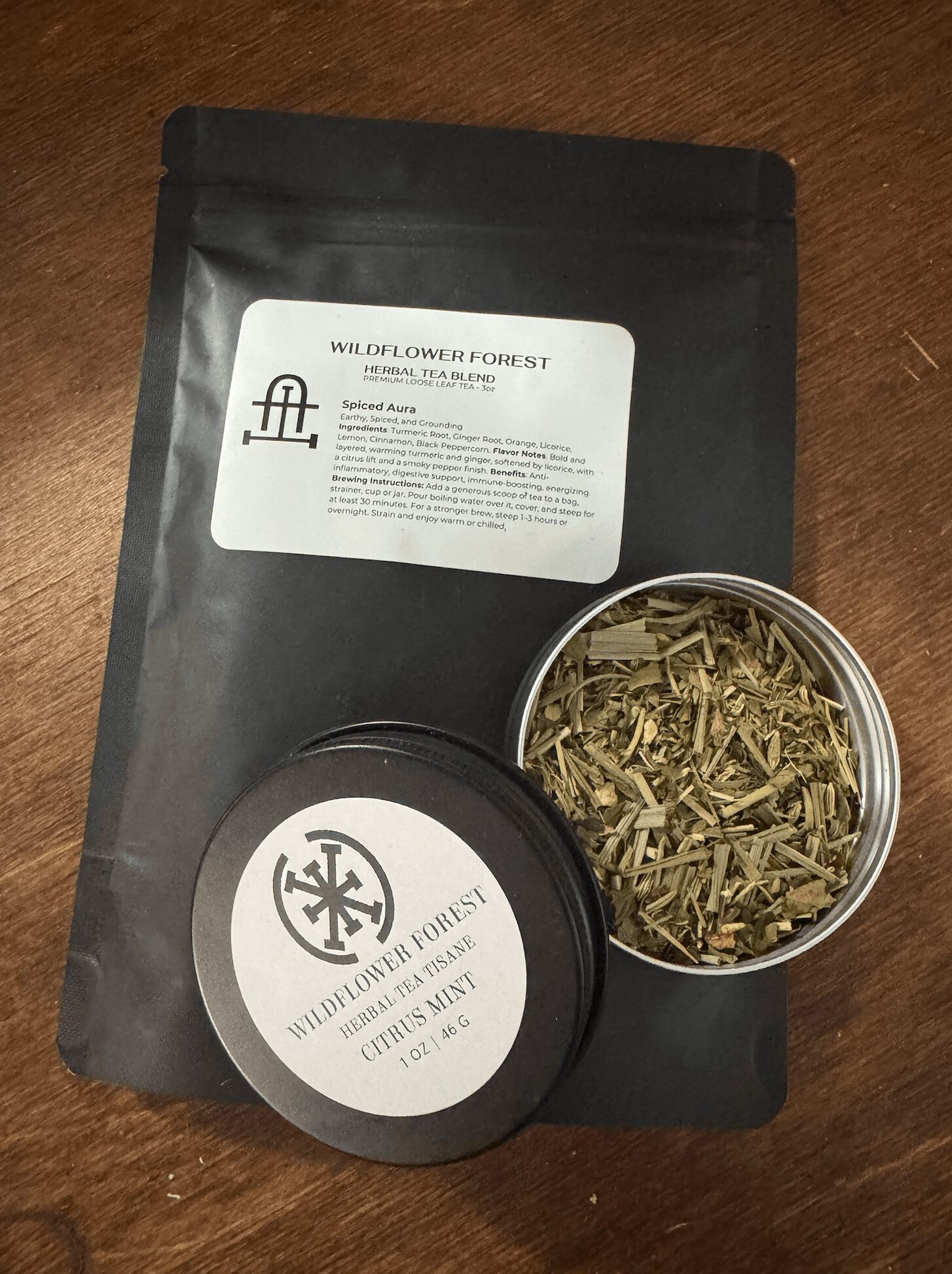 Black resealable bag with a white label, open round container with dried herbs, and a black lid with a logo, on wooden surface.