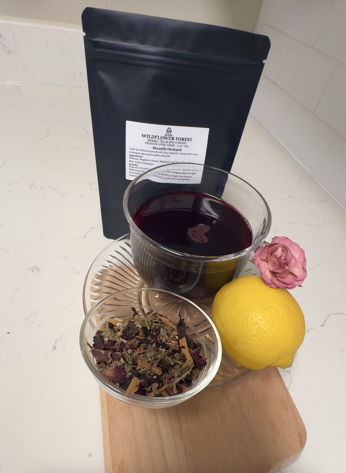 Glass of dark red liquid, lemon, pink flower, herbal tea in a glass bowl, black package in background, on wooden surface.