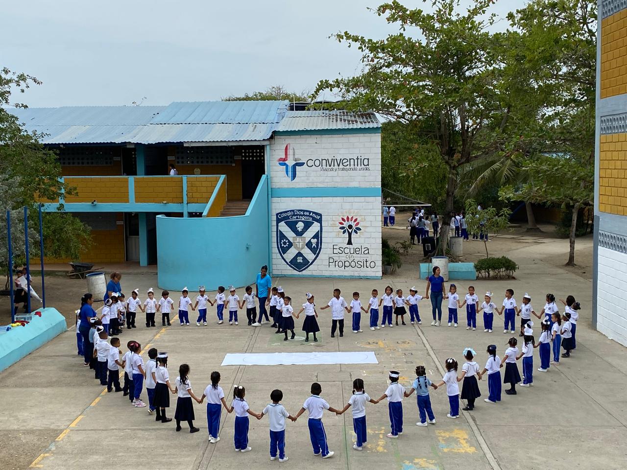 Students form a large circle outside a school building, holding hands, with trees and a sign in the background.