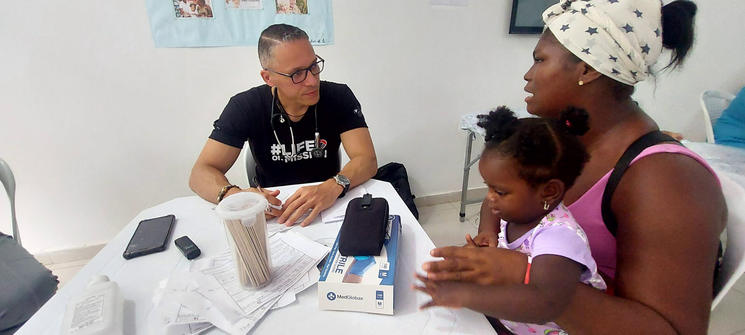 A woman and a young girl sit at a table with a man across from them, in a room with white walls.