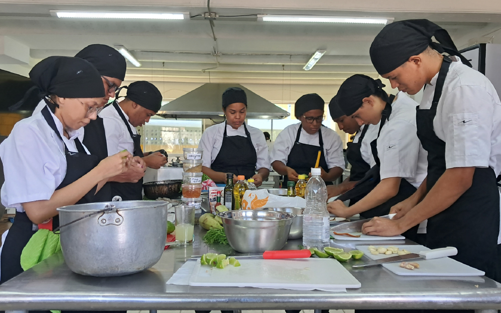 Group of people in chef uniforms and black head coverings preparing food in a kitchen.