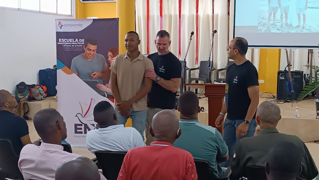 Three men stand in front of a seated audience in a room with a projector screen and a banner, engaging in a presentation.