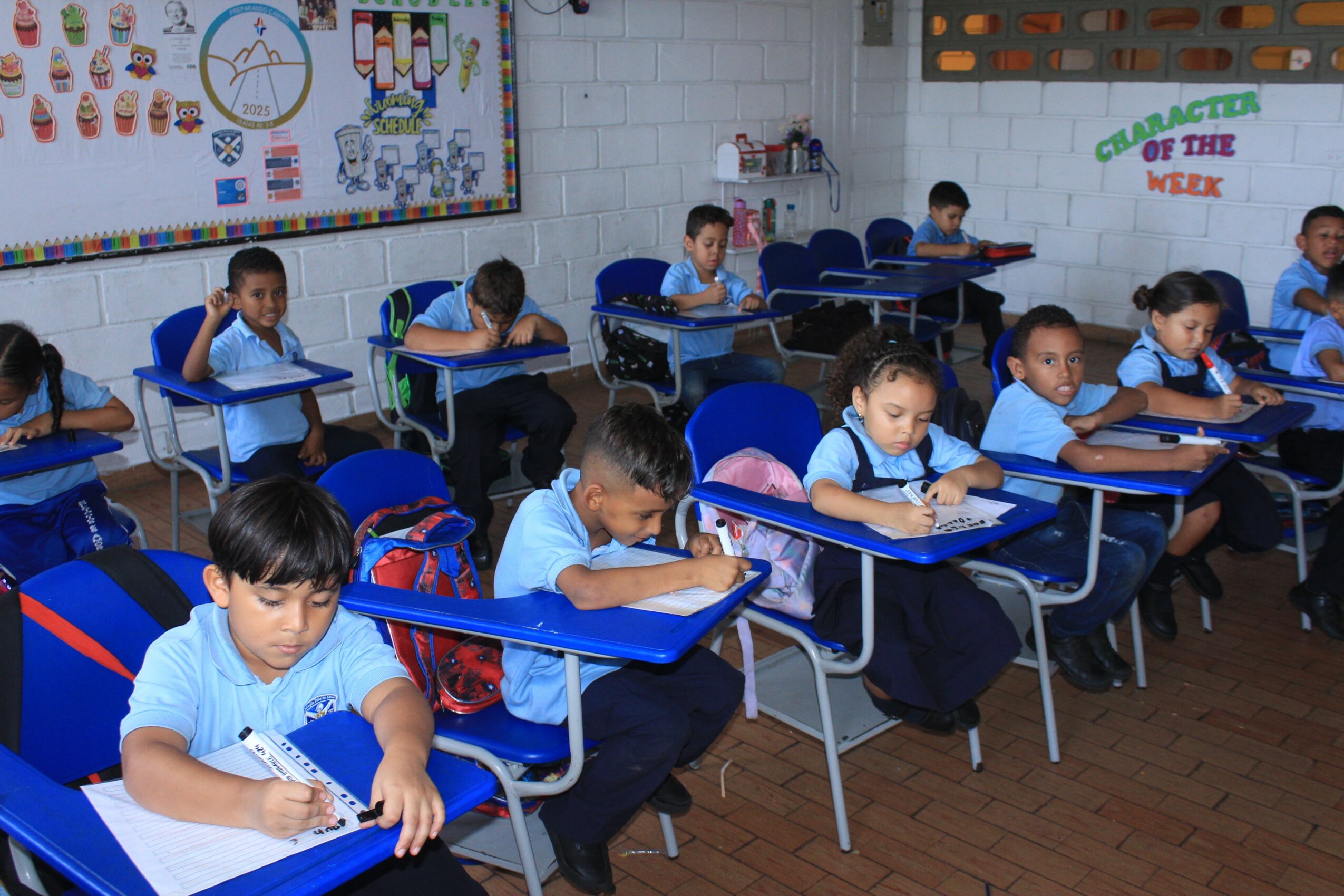 Classroom with young students sitting at desks, writing, wearing blue uniforms, and backpacks on chairs, white brick wall background.