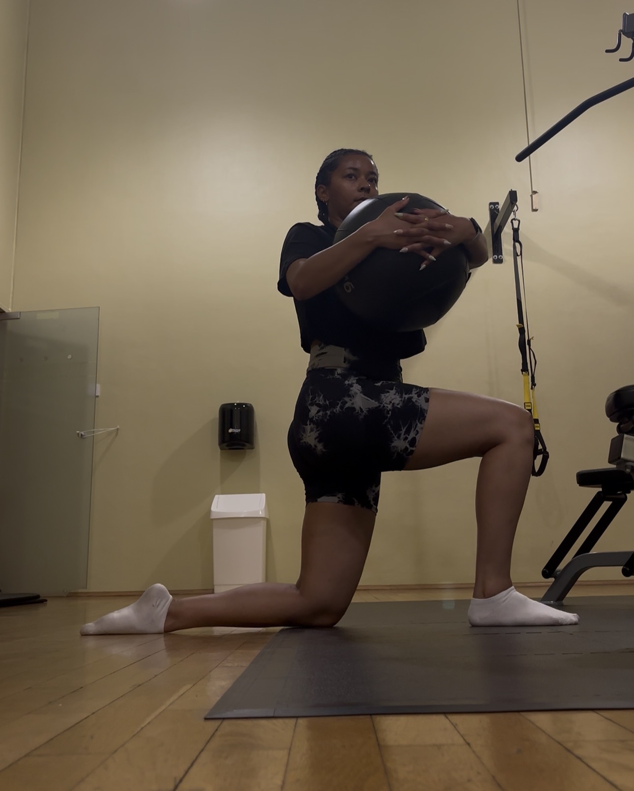 Person in workout attire holding a medicine ball in a gym, kneeling on one knee on a mat.