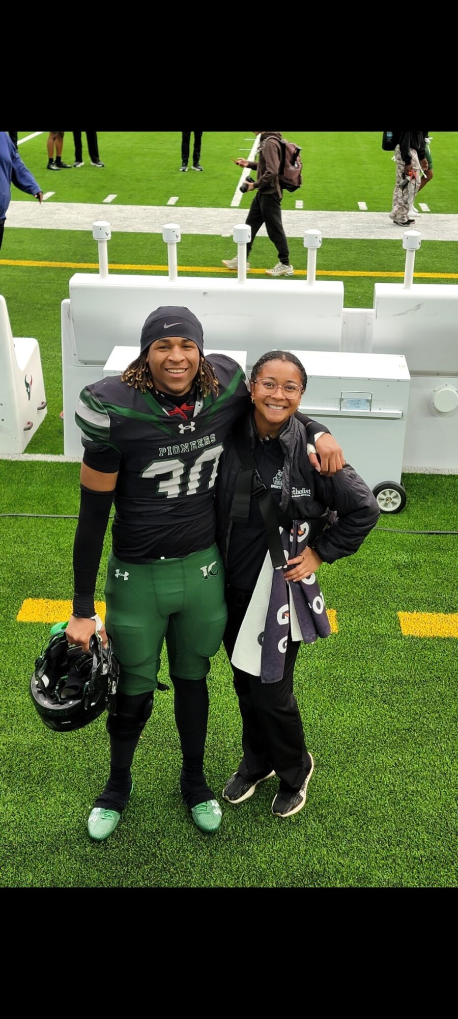 Two people in football uniforms standing on a field, smiling, with benches and other people in the background.