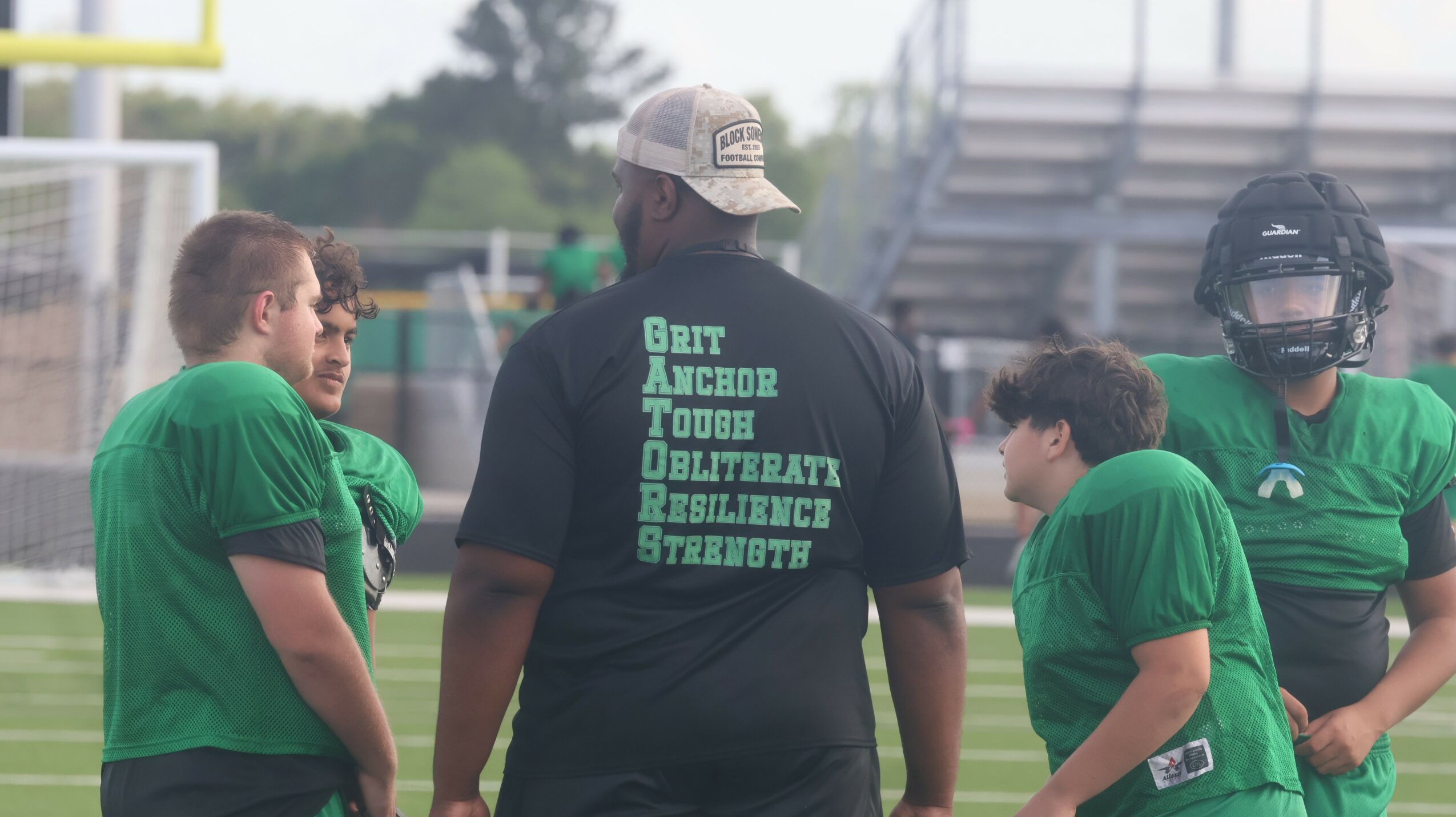 Four football players in green uniforms and a coach in black shirt on field, with bleachers in background.