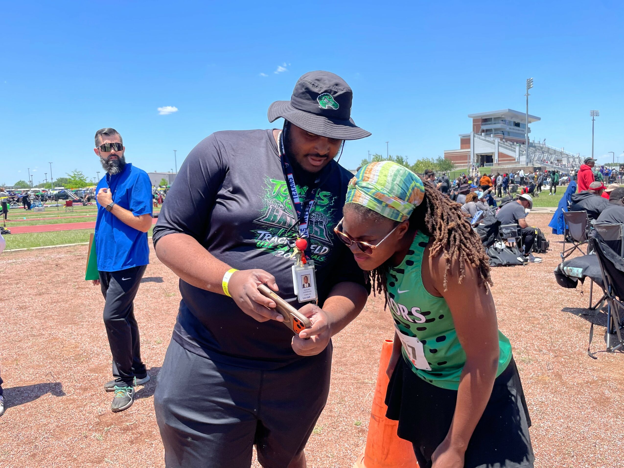 Two women look at a phone together at an outdoor event, with a man in a blue shirt in the background, on a sunny day.