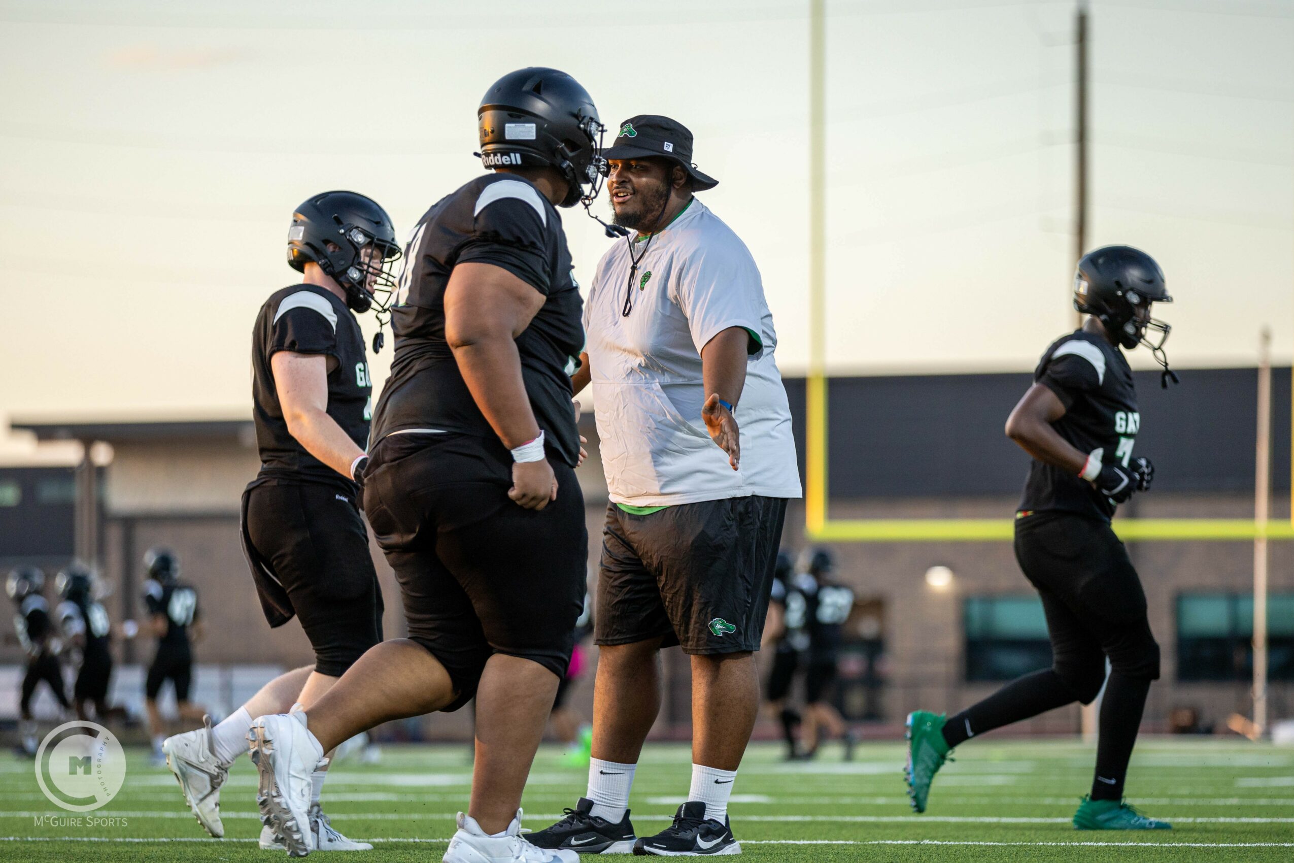 Football coach talking to players on field during practice, players wearing helmets and athletic gear, stadium in background