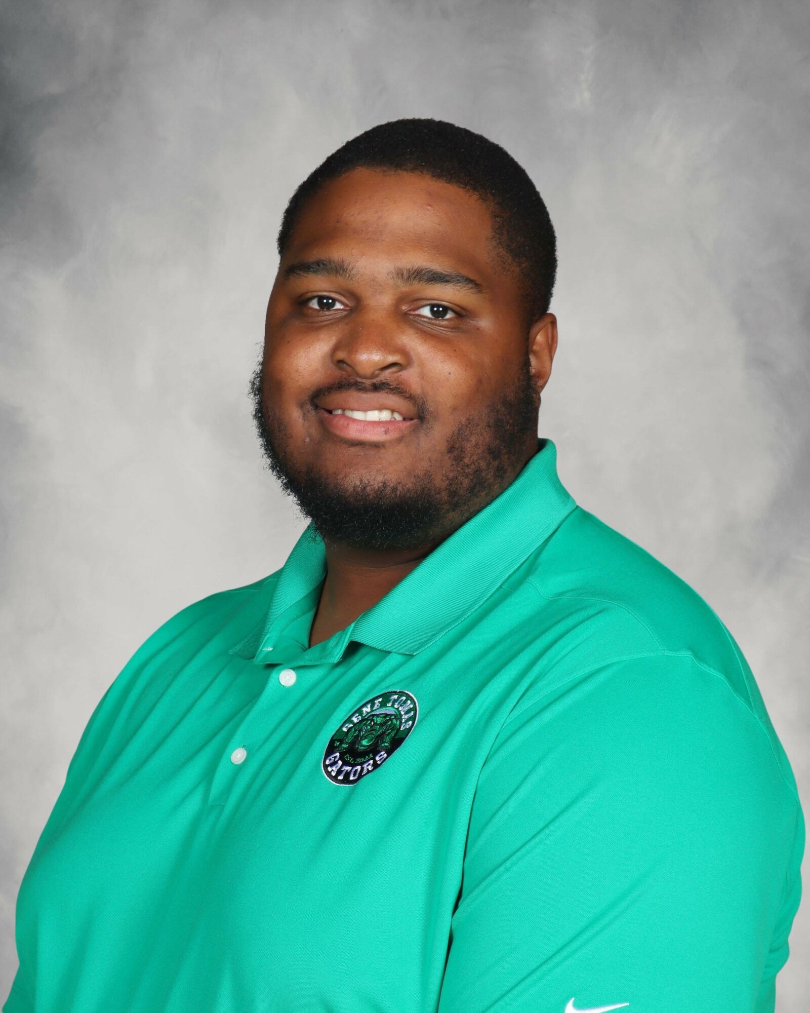 Portrait of a man with short hair and beard wearing a teal polo shirt with a logo, smiling, against a gray background.