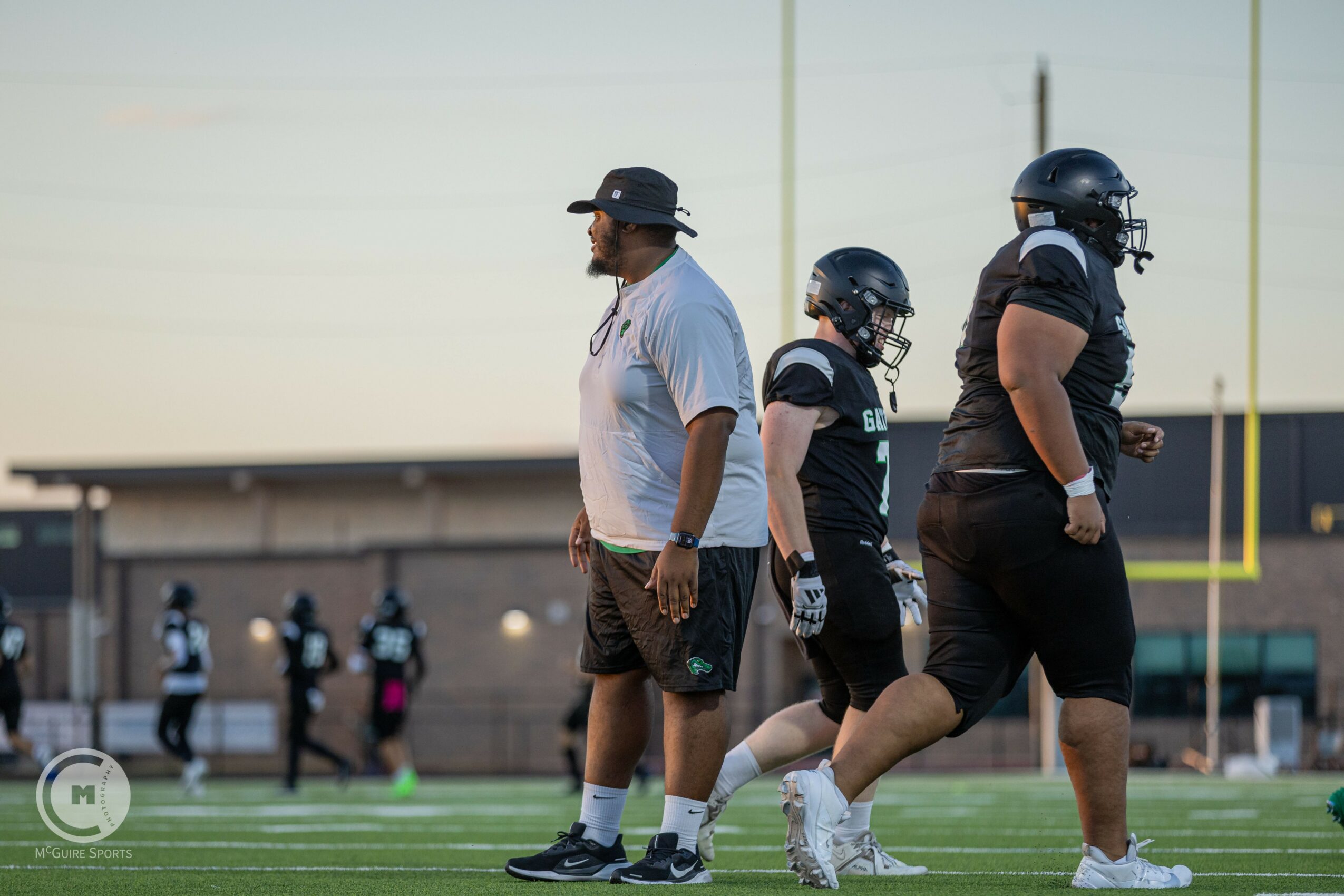 Football coach and players on field during practice, some wearing helmets, others walking or standing, background shows goalpost and field.