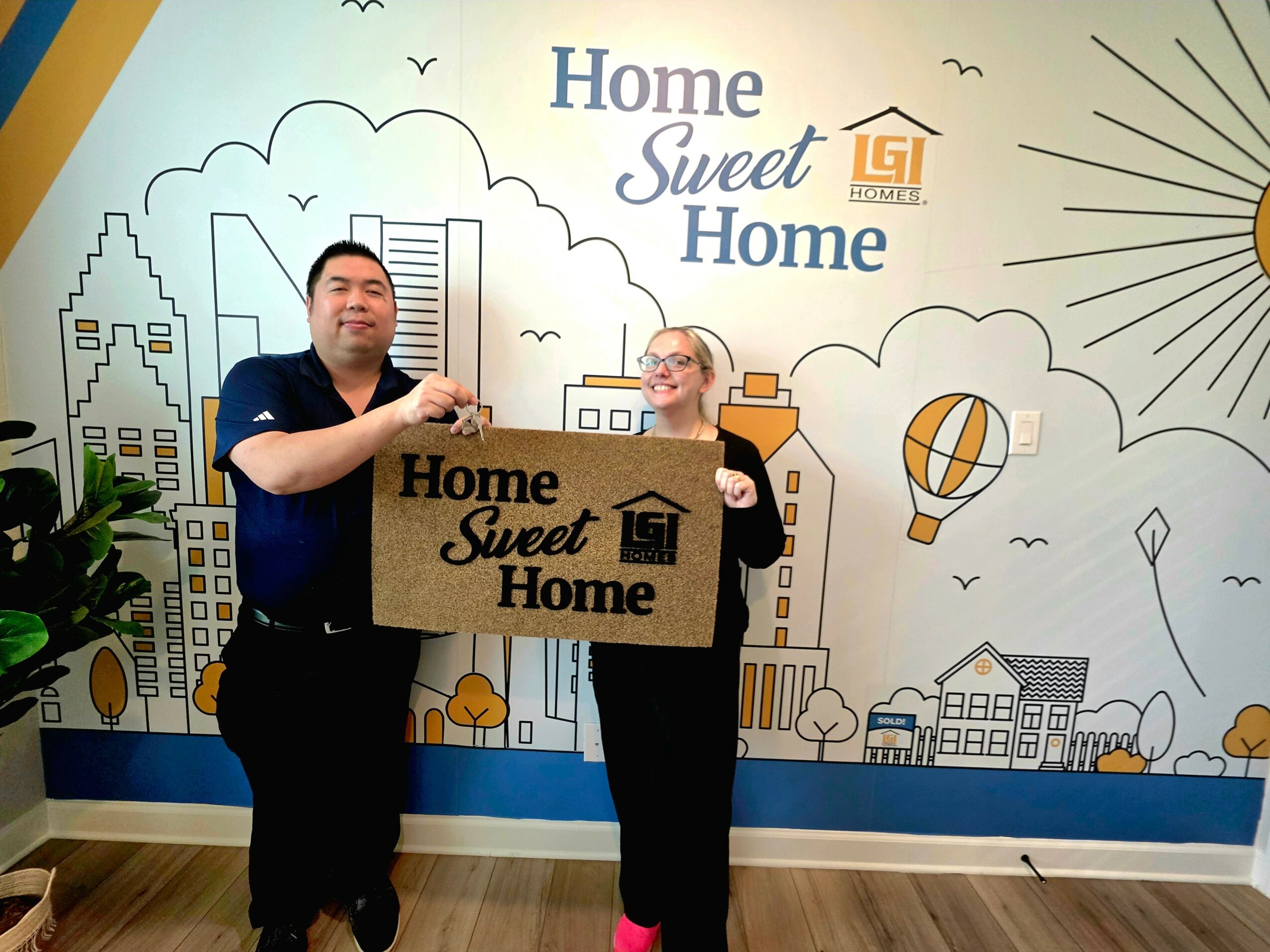 Two people holding a sign that says 'Home Sweet Home' in front of a colorful cityscape mural.