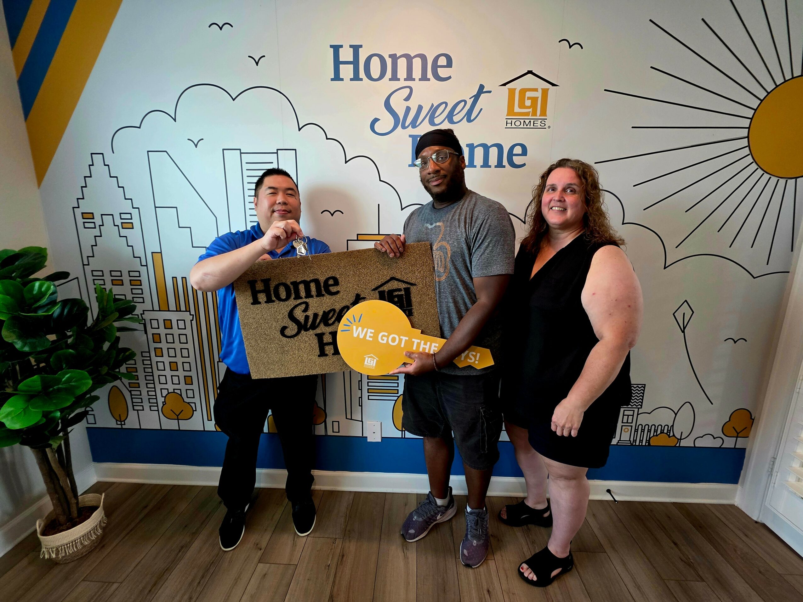 Three people standing in front of a wall with a cityscape and sun illustration, holding a large key and a sign, smiling.