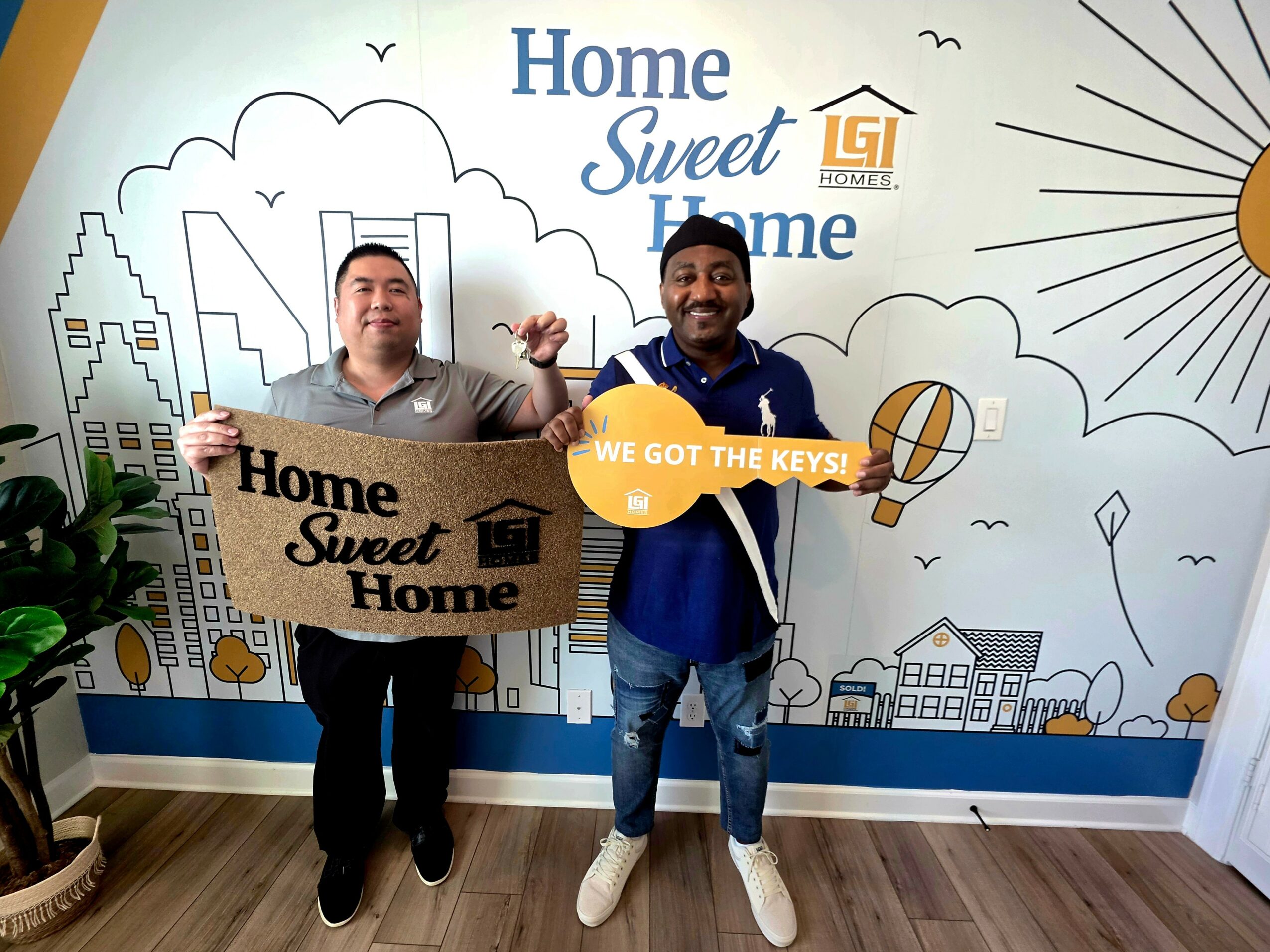 Two men standing in front of a wall with a cityscape and sun illustration, holding a sign that says 'Home Sweet Home' and a key, smiling.