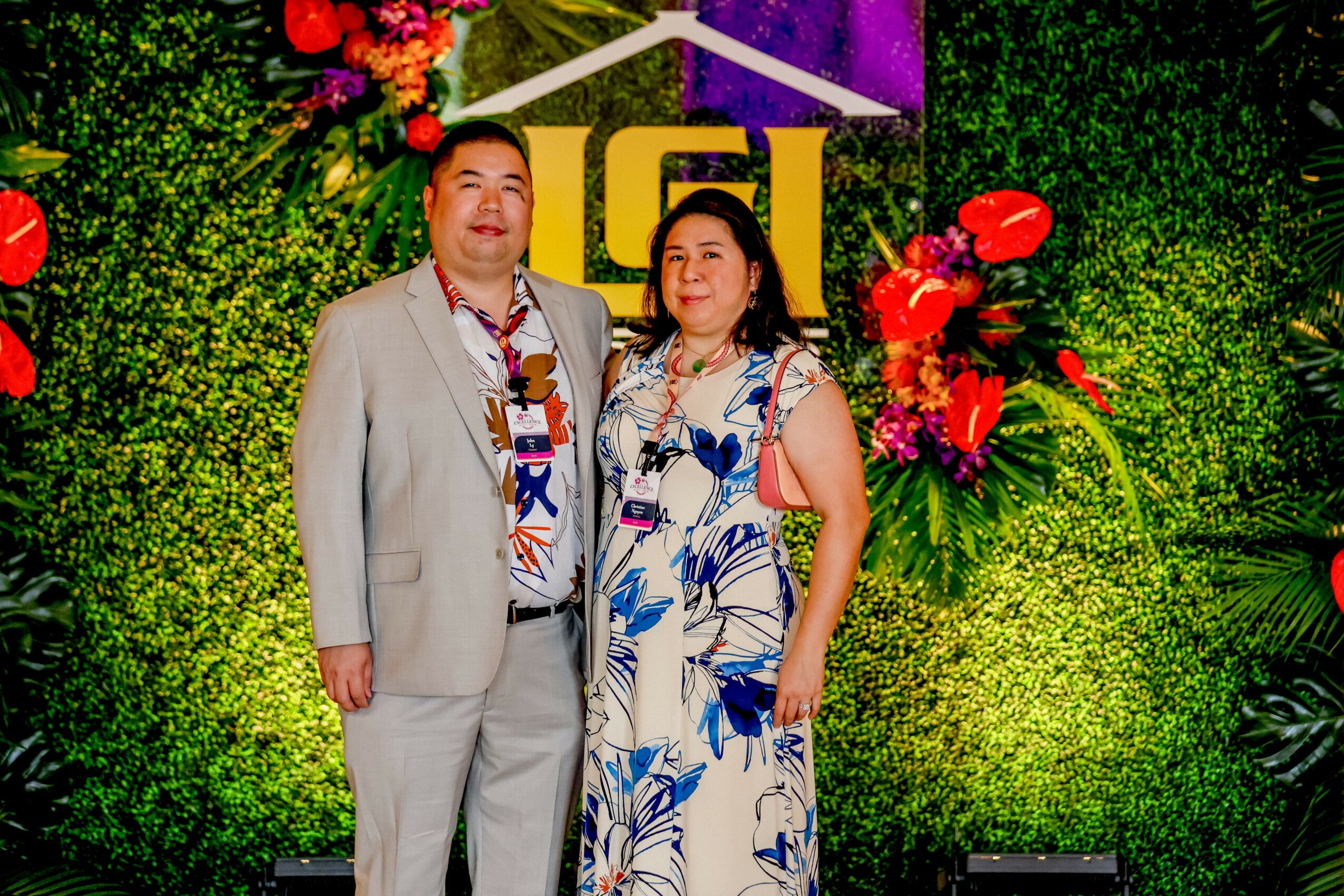 A man and woman stand together in front of a decorated green backdrop with flowers, posing for a photo.