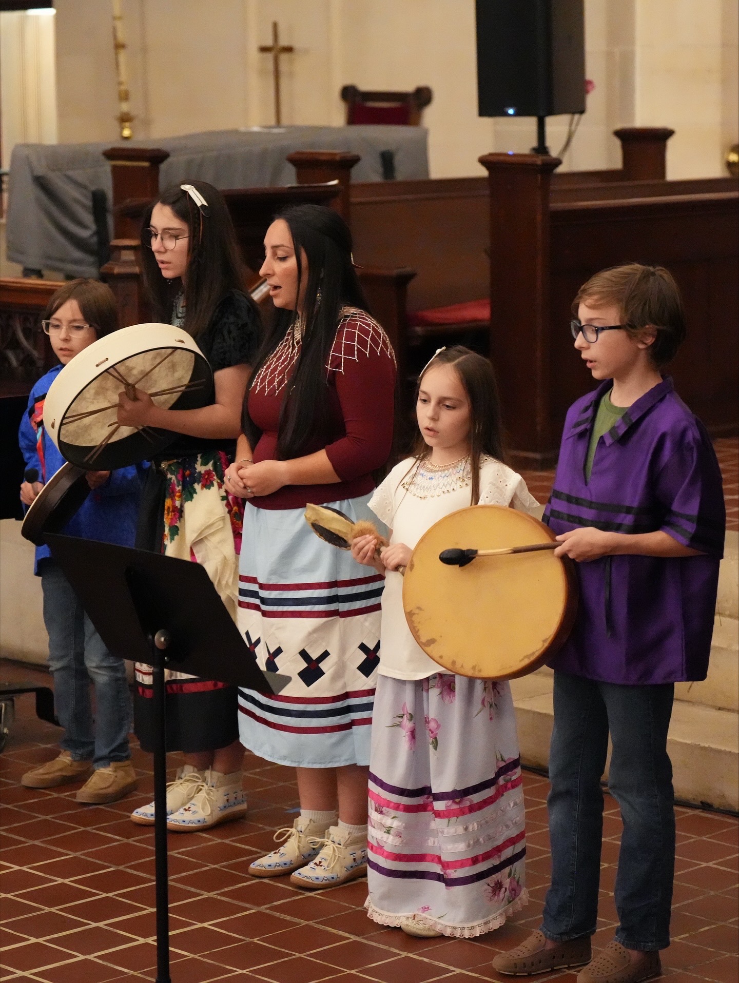 Group of children and young women holding musical instruments in a church setting, standing in a row, some reading music sheets.