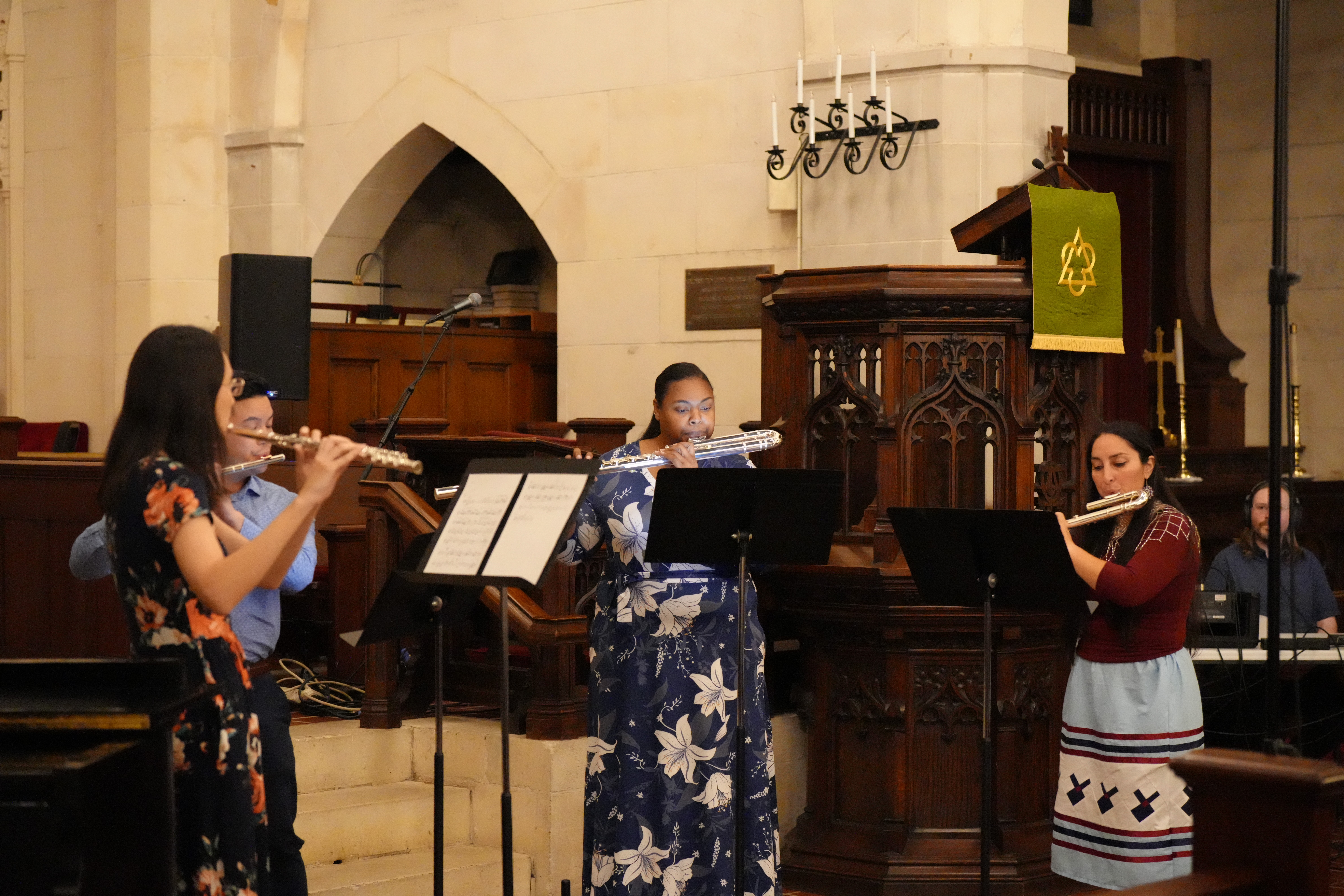 Three women playing flutes in a church with wooden pulpit and arched architecture, wearing colorful dresses, with sheet music stands.