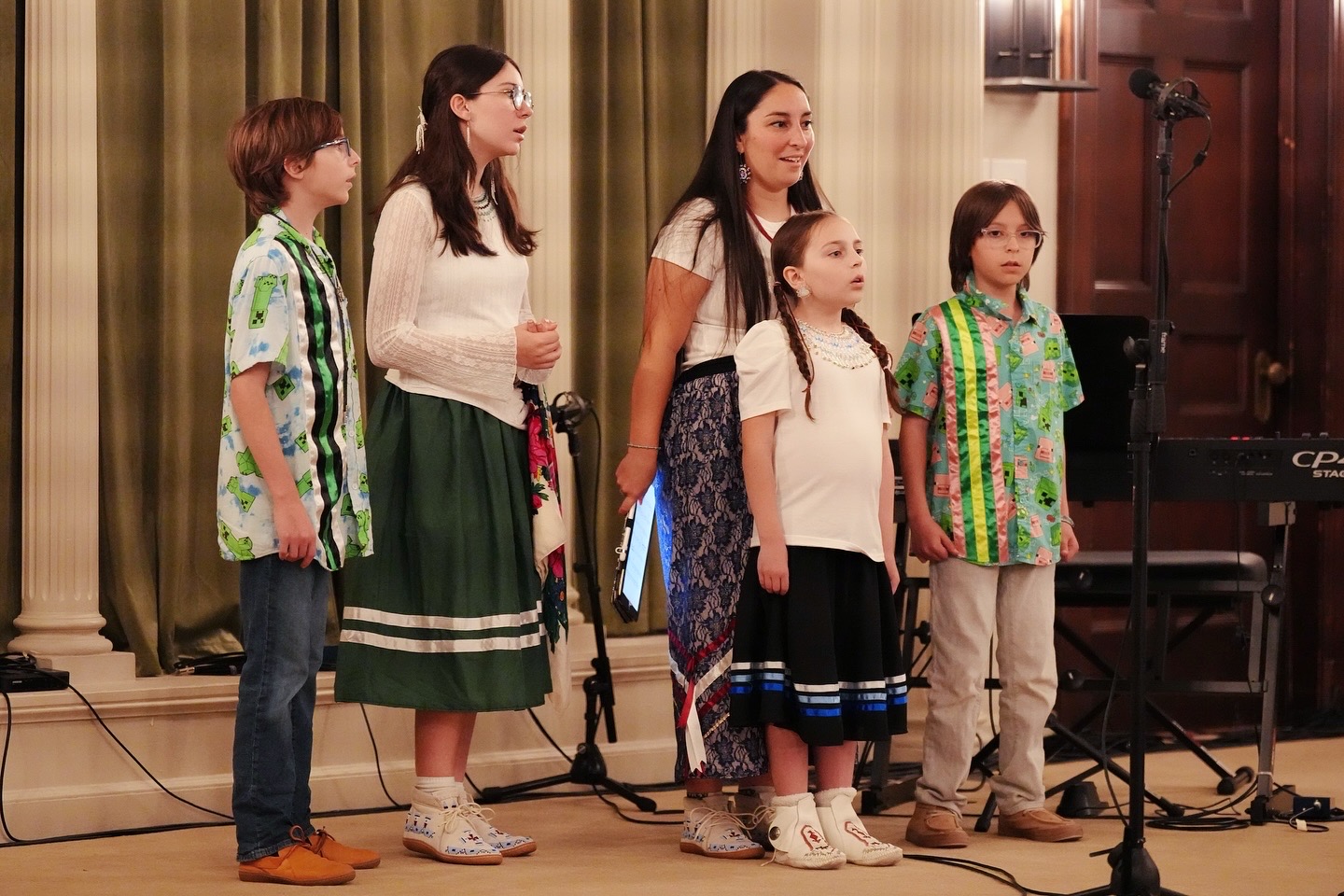 Group of six children standing on stage, smiling, with a curtain background and microphones nearby.