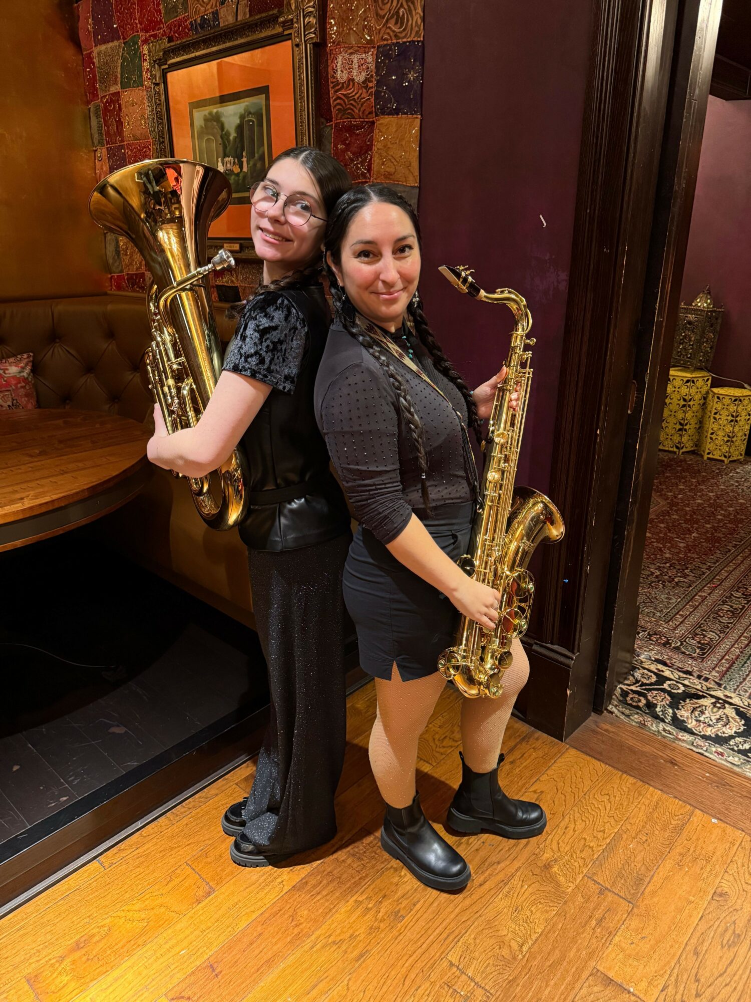 Two women holding saxophones and smiling, standing indoors with wooden flooring and decorative wall elements.