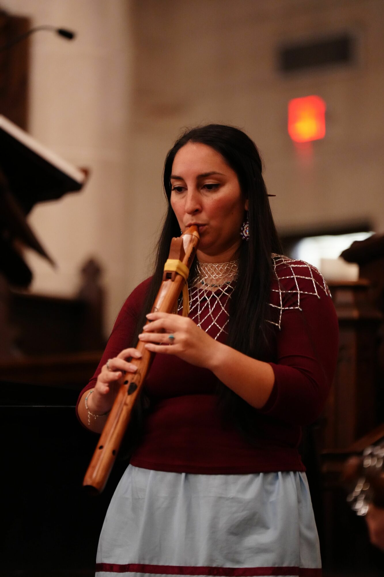 Woman playing a flute indoors, with a blurred background and a red exit sign visible.