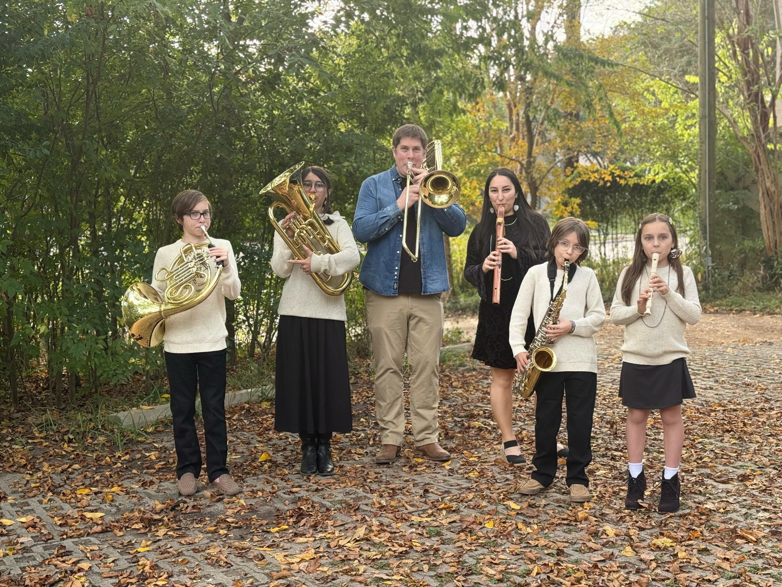 Group of six people playing musical instruments outdoors in a park with trees and fallen leaves.