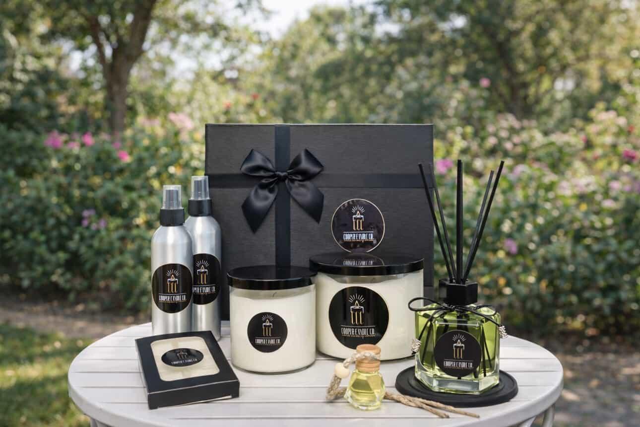 Gift set with black box, candles, sprays, and reed diffuser on a white table outdoors, greenery in background.