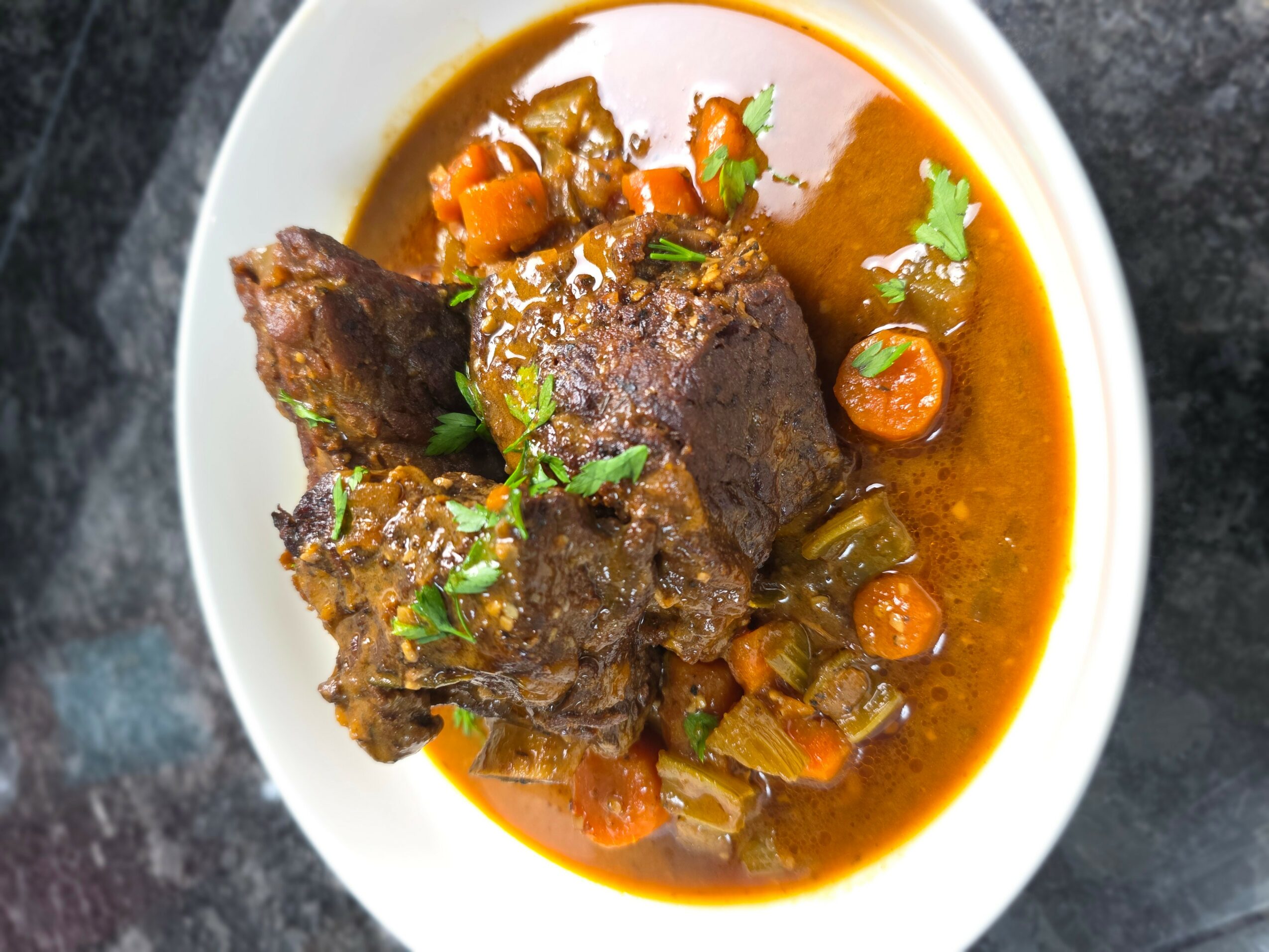 Bowl of beef stew with carrots, celery, and herbs in a reddish broth, served in a white bowl on a dark surface.