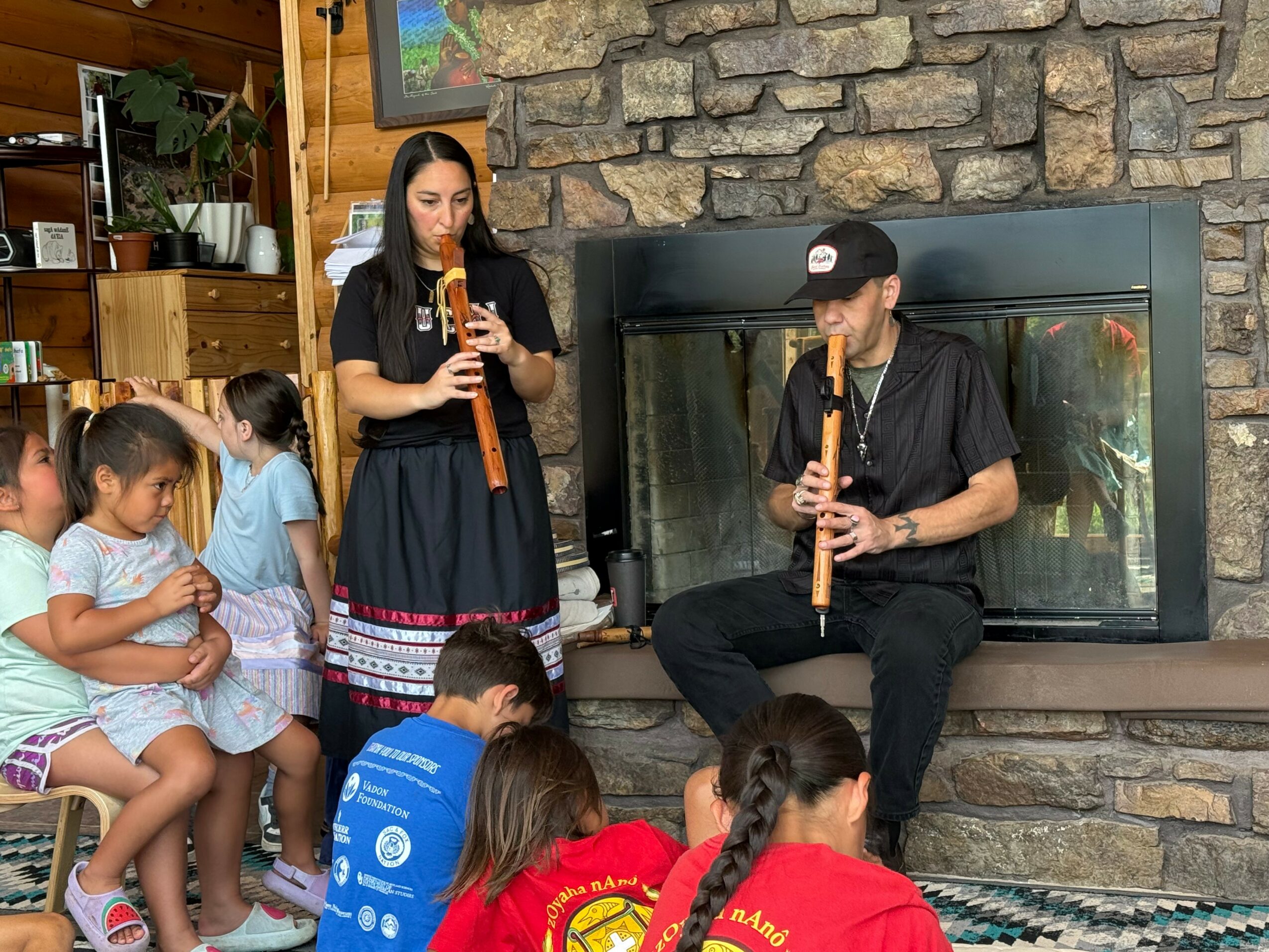 Two musicians playing flutes in front of children sitting on a bench, with a stone fireplace and wooden wall in background.