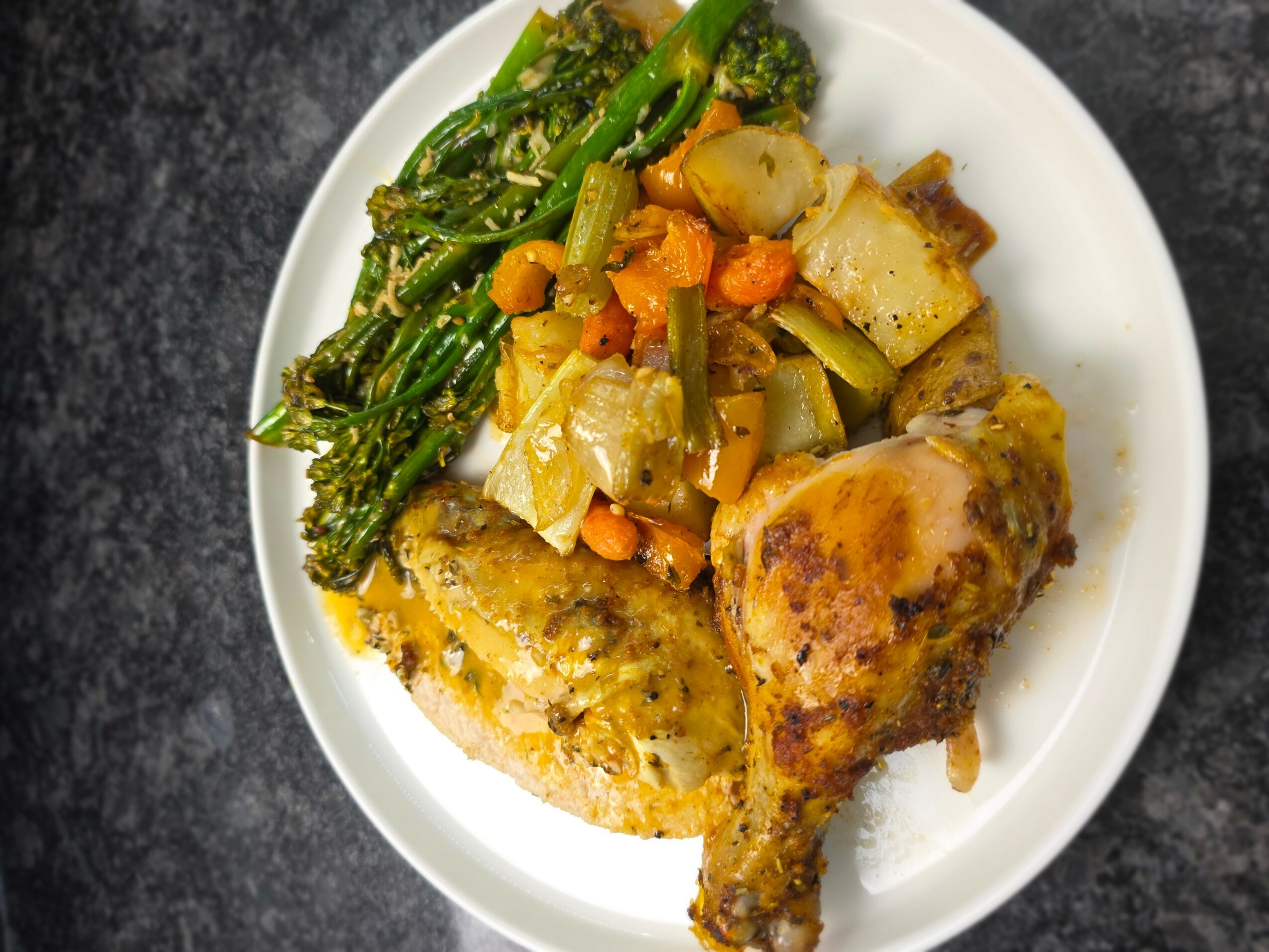 Plate with cooked chicken, mixed vegetables including carrots, potatoes, and greens, on a dark surface.