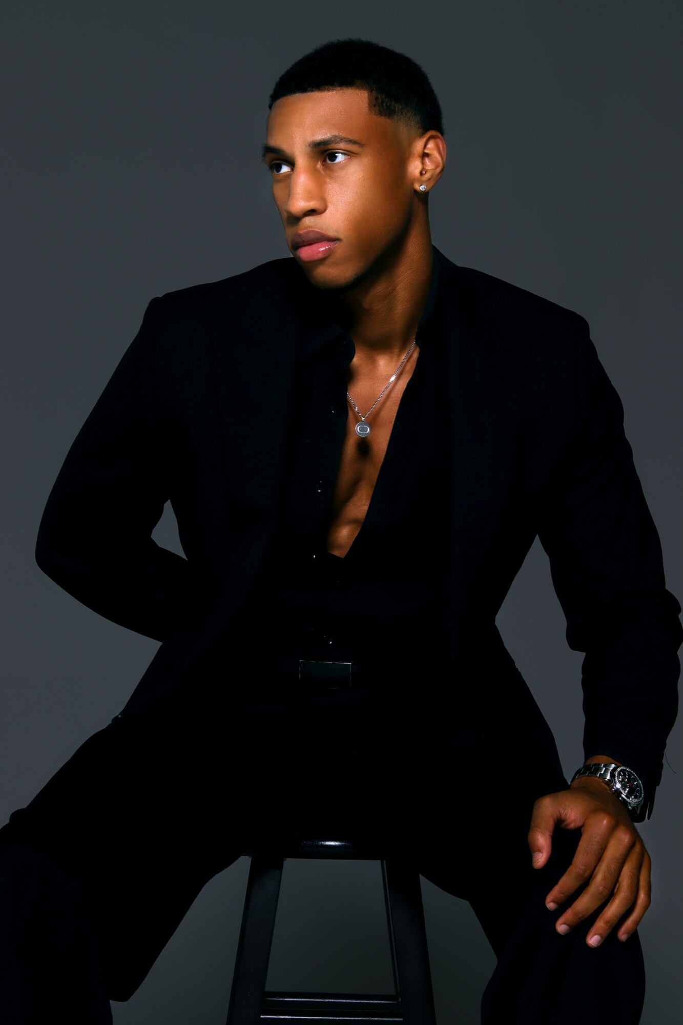 Young man with short hair wearing a black blazer and shirt, sitting against a dark background.