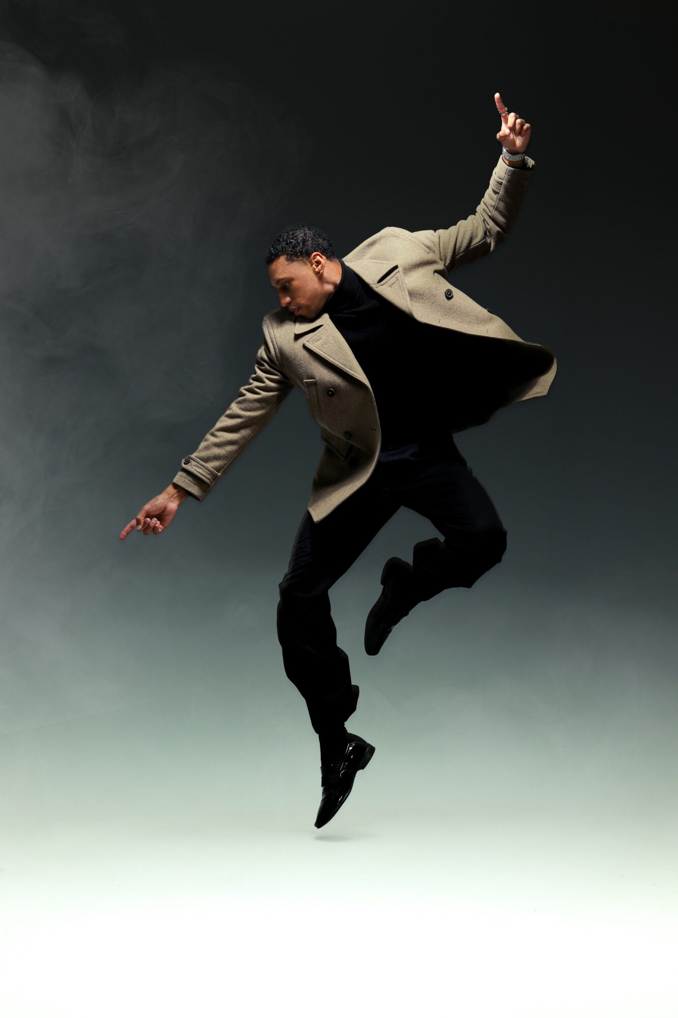 Person mid-air with arms raised, wearing a beige coat and black pants, against a dark background.