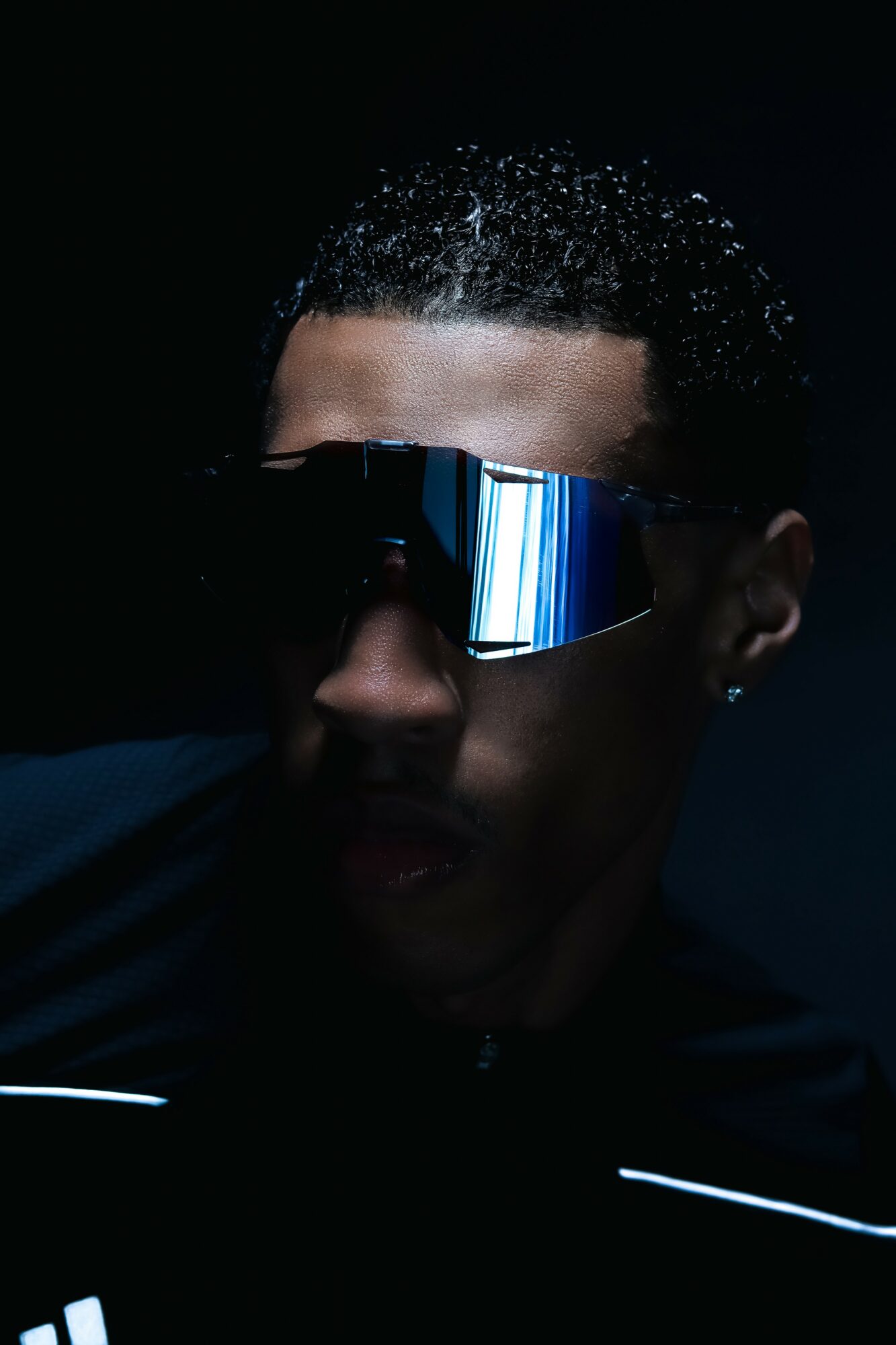 Person wearing reflective sunglasses with short curly hair, dark background, and minimal lighting, facing forward.
