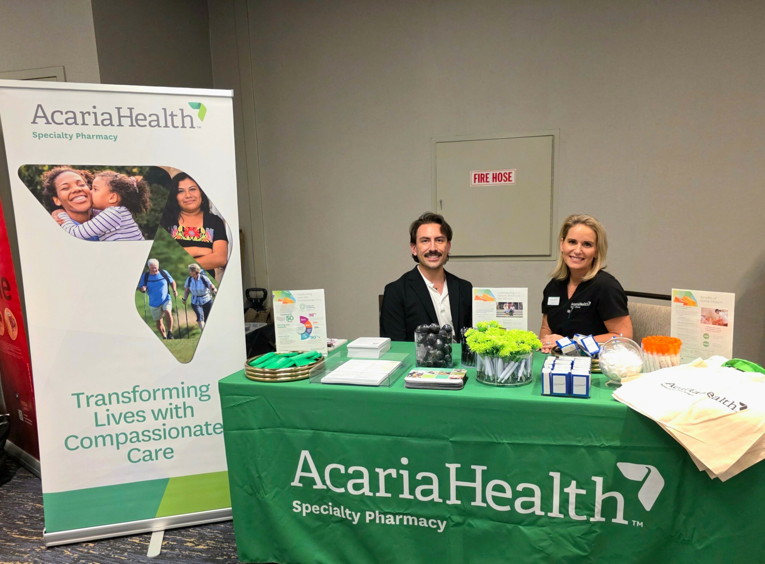 Two people sit behind a table with AcariaHealth branding, promotional materials, and a banner, in an indoor setting.