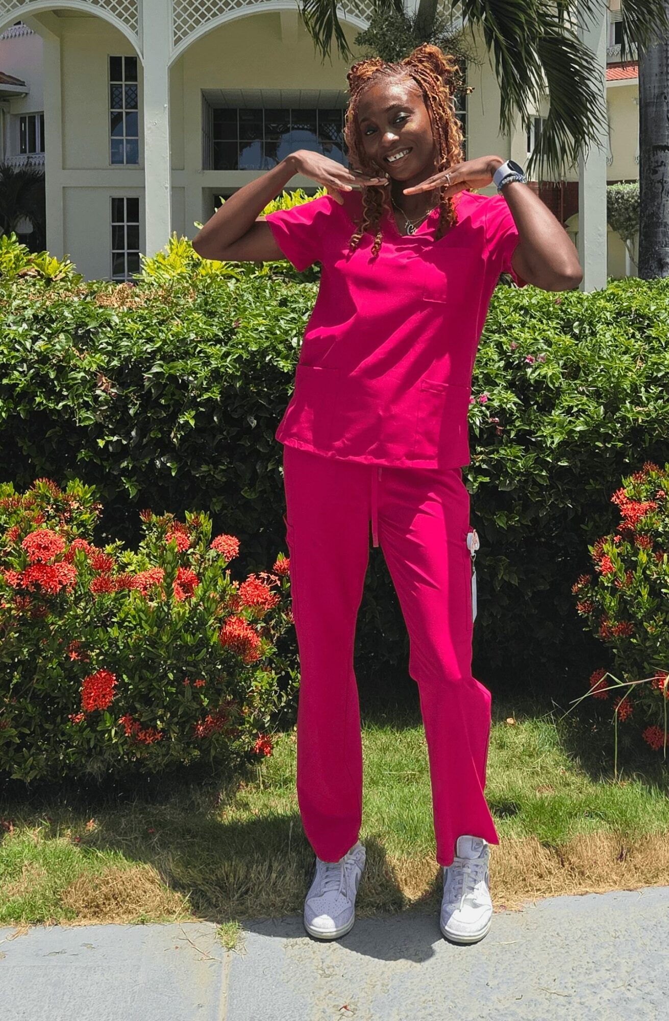 Woman in pink scrubs standing outdoors with bushes and a building in background, smiling and posing with arms raised.