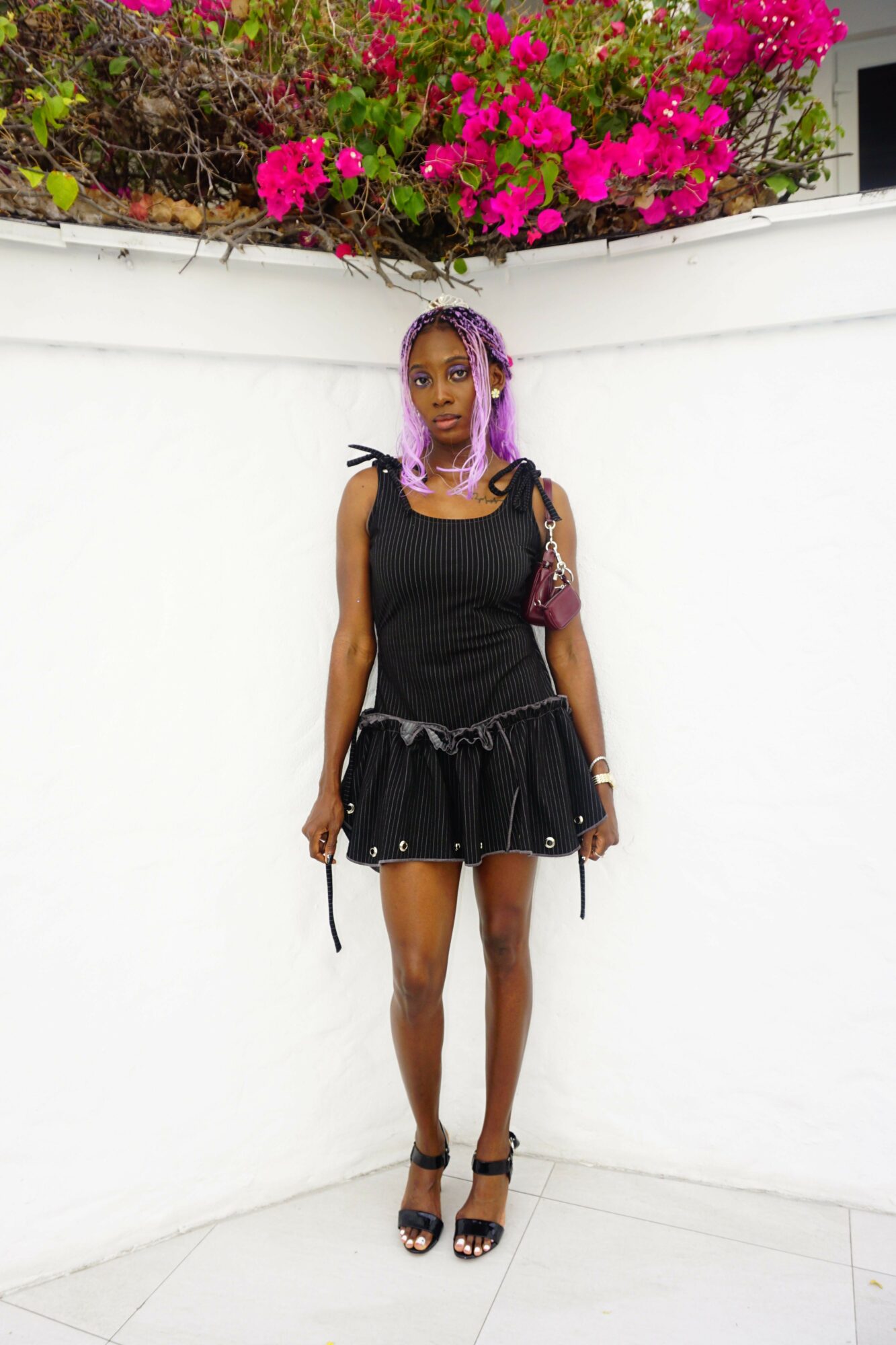 Woman with purple hair in a black dress standing against a white wall with pink flowers, smiling.