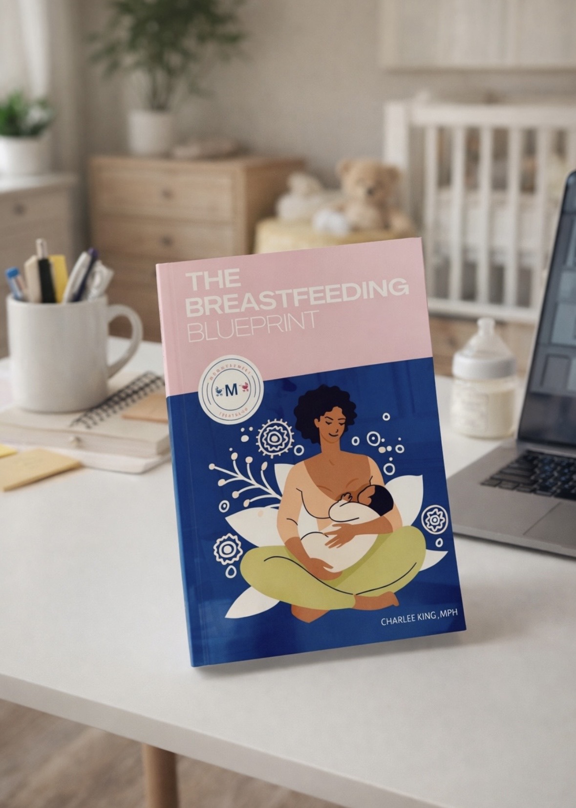 Book titled 'The Breastfeeding Experiment' on a desk with a laptop, pen holder, and background of a nursery room.