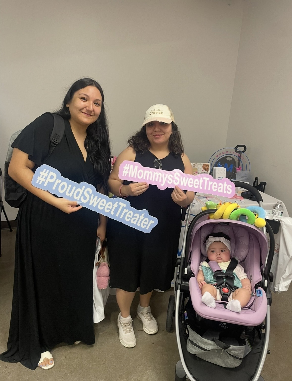Two women and a baby in a stroller indoors, holding signs that read #ProudSweetTreater and #MommySweetTreats.
