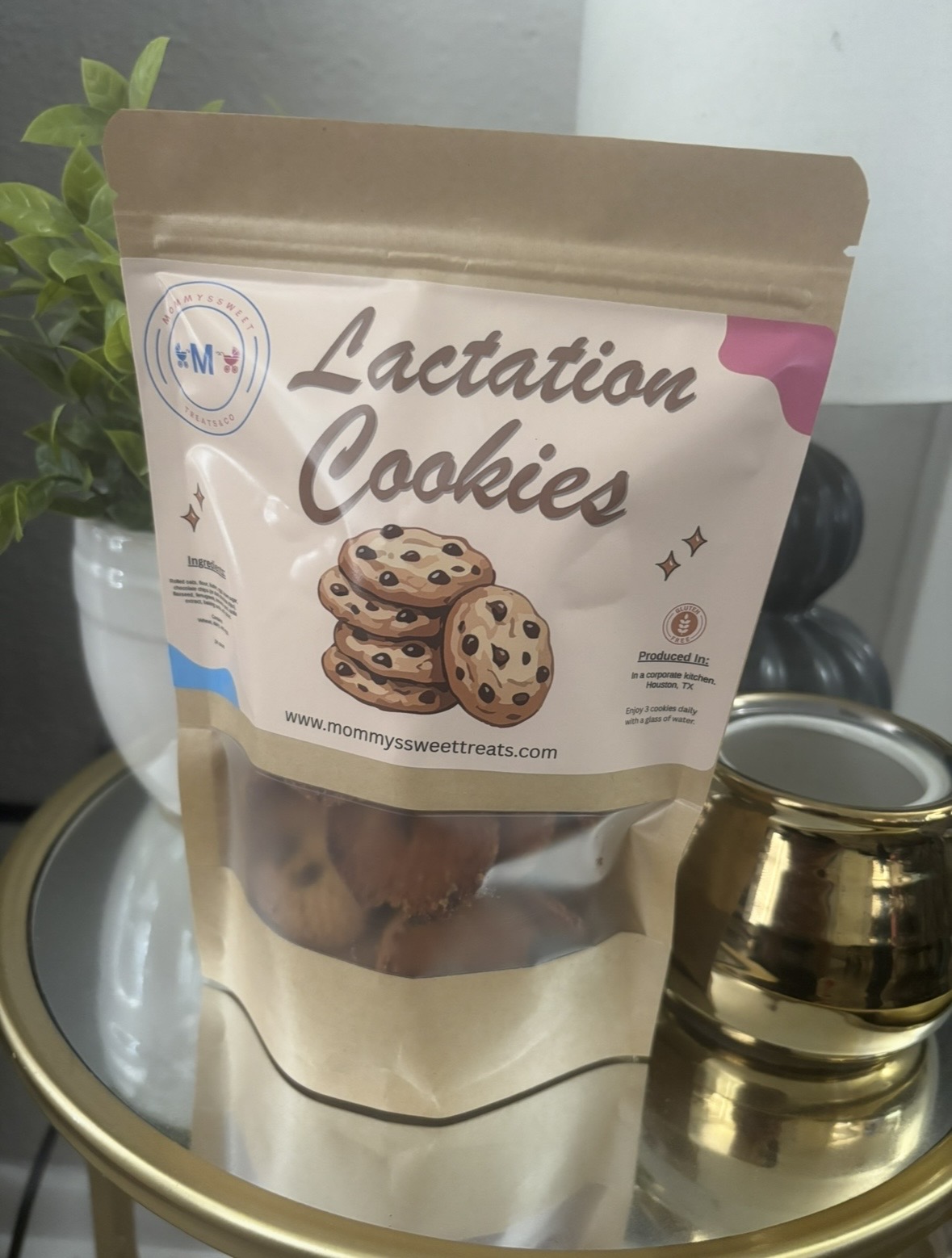Bag of Lactation Cookies with cookies illustration, placed on a reflective surface with a plant and a gold container nearby.