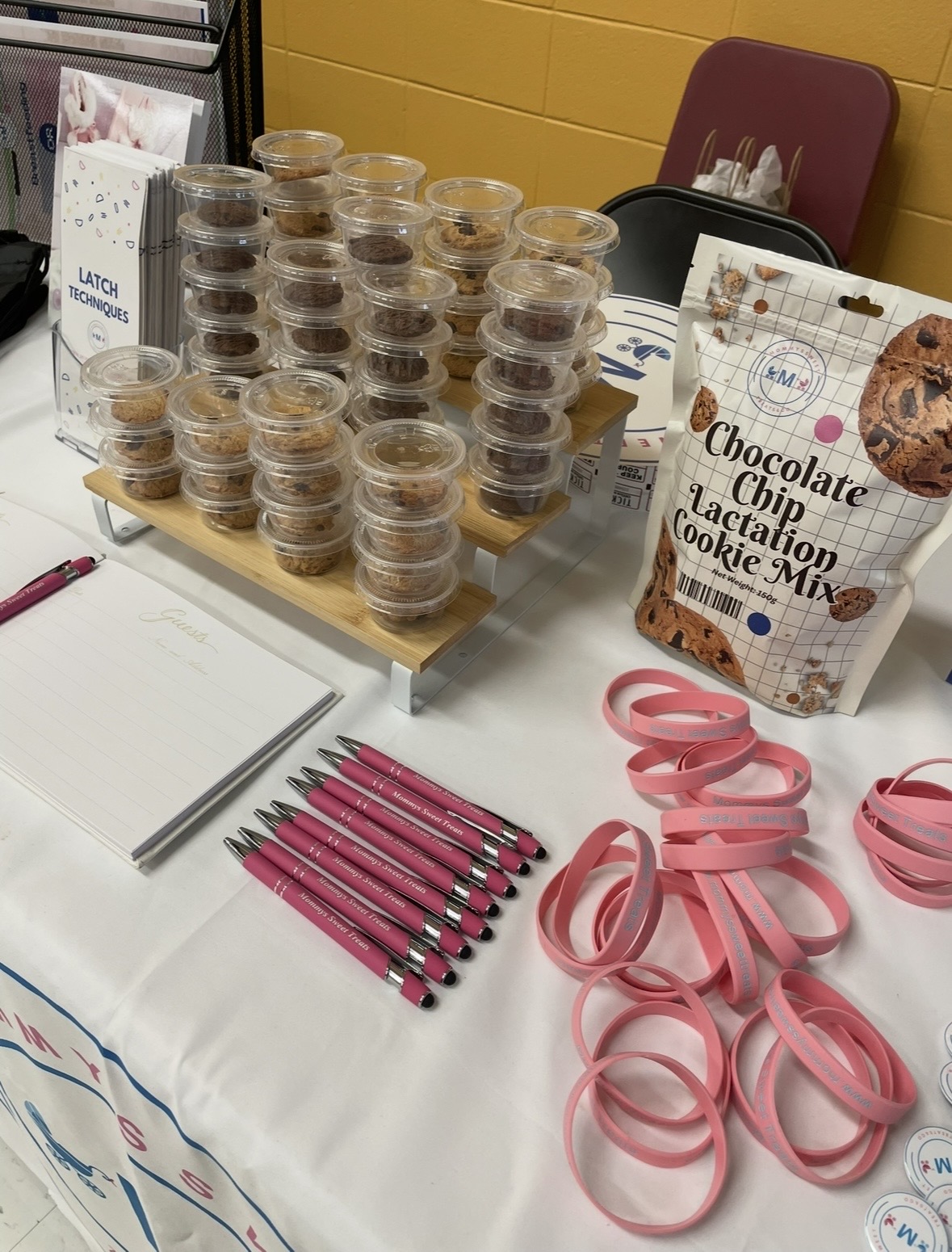 Table with stacked small containers, a bag of chocolate chip cookie mix, pink ribbons, and pens, against a yellow wall.