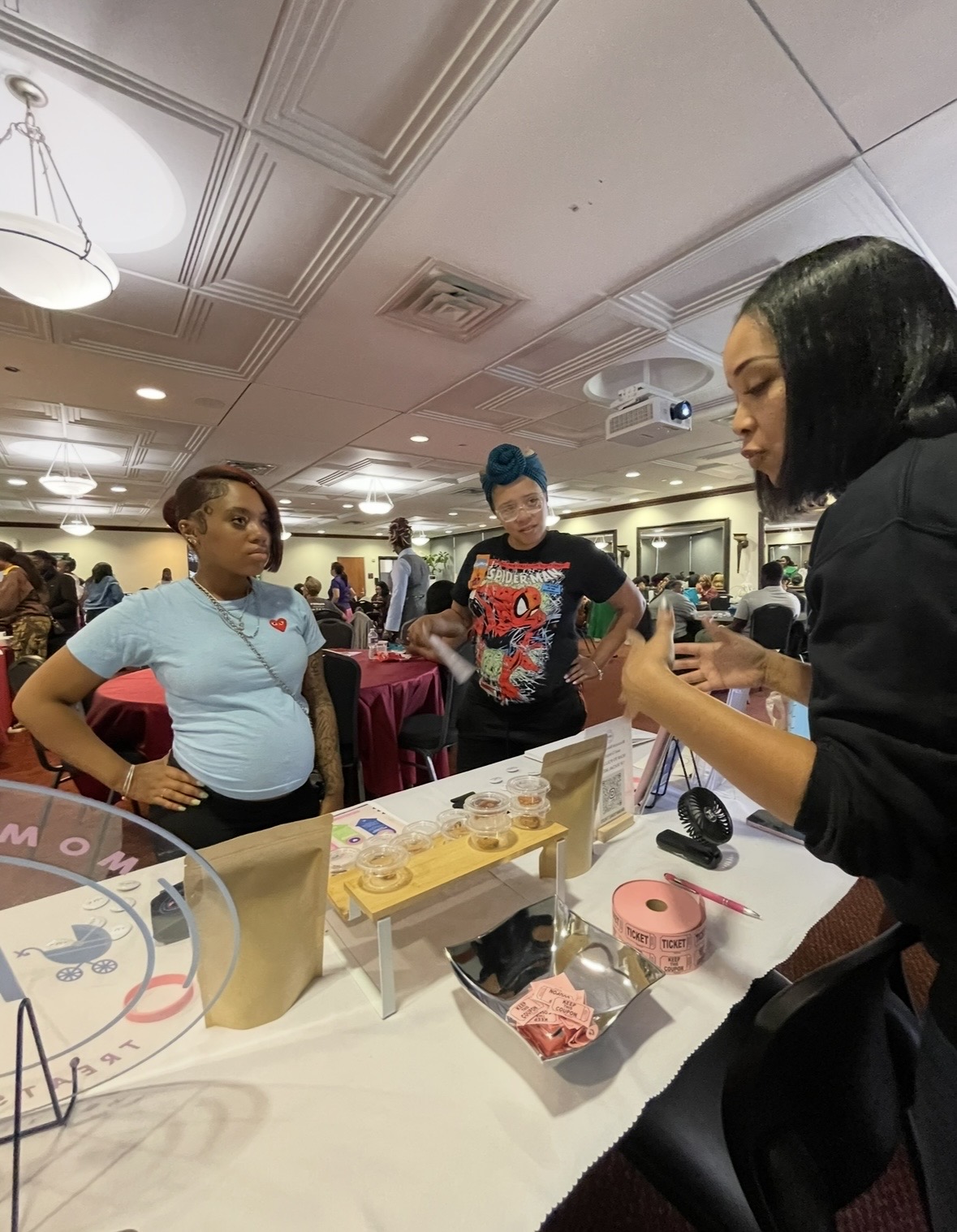 Woman with black hair demonstrating a product at a booth, with two women observing in a crowded indoor event space.