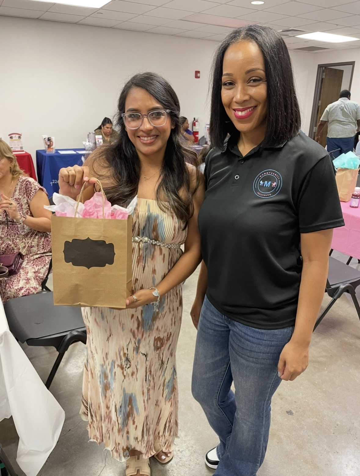 Two women smiling, one holding a gift bag, in a room with other people and tables.