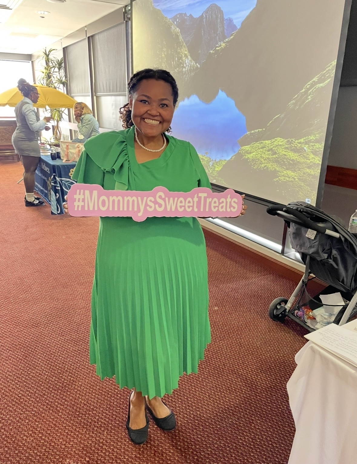Woman in green dress holding a pink sign with hashtag #MommysSweetTreats, smiling indoors with a scenic background projection.