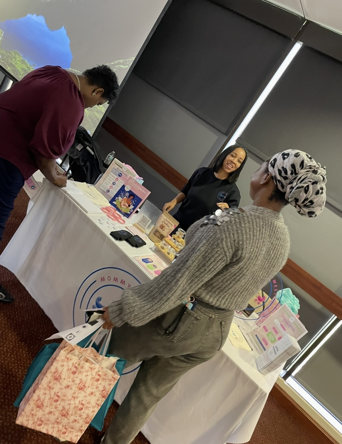 Three women at an indoor event table with promotional materials, one woman wearing a headscarf, another smiling, and a third looking at items.